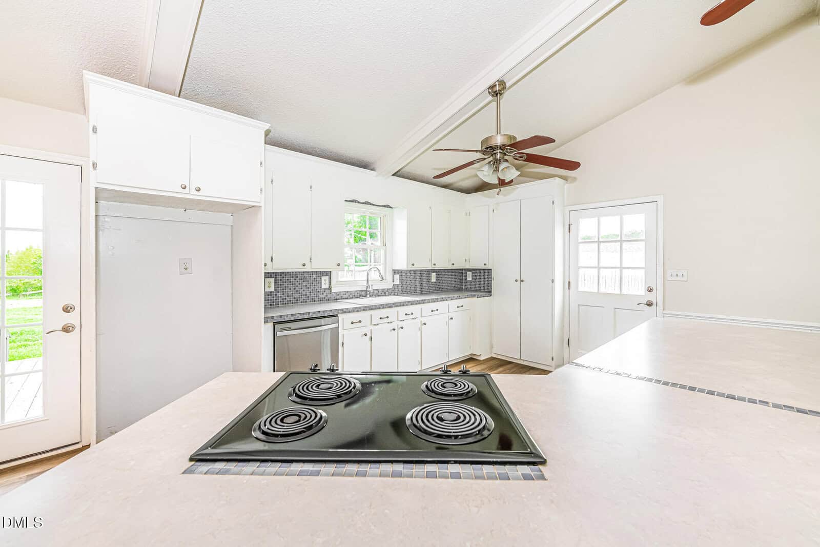 Bright kitchen with white cabinets, gray tile backsplash, ceiling fan, and electric stovetop on a light countertop; two doors and a window provide natural light.