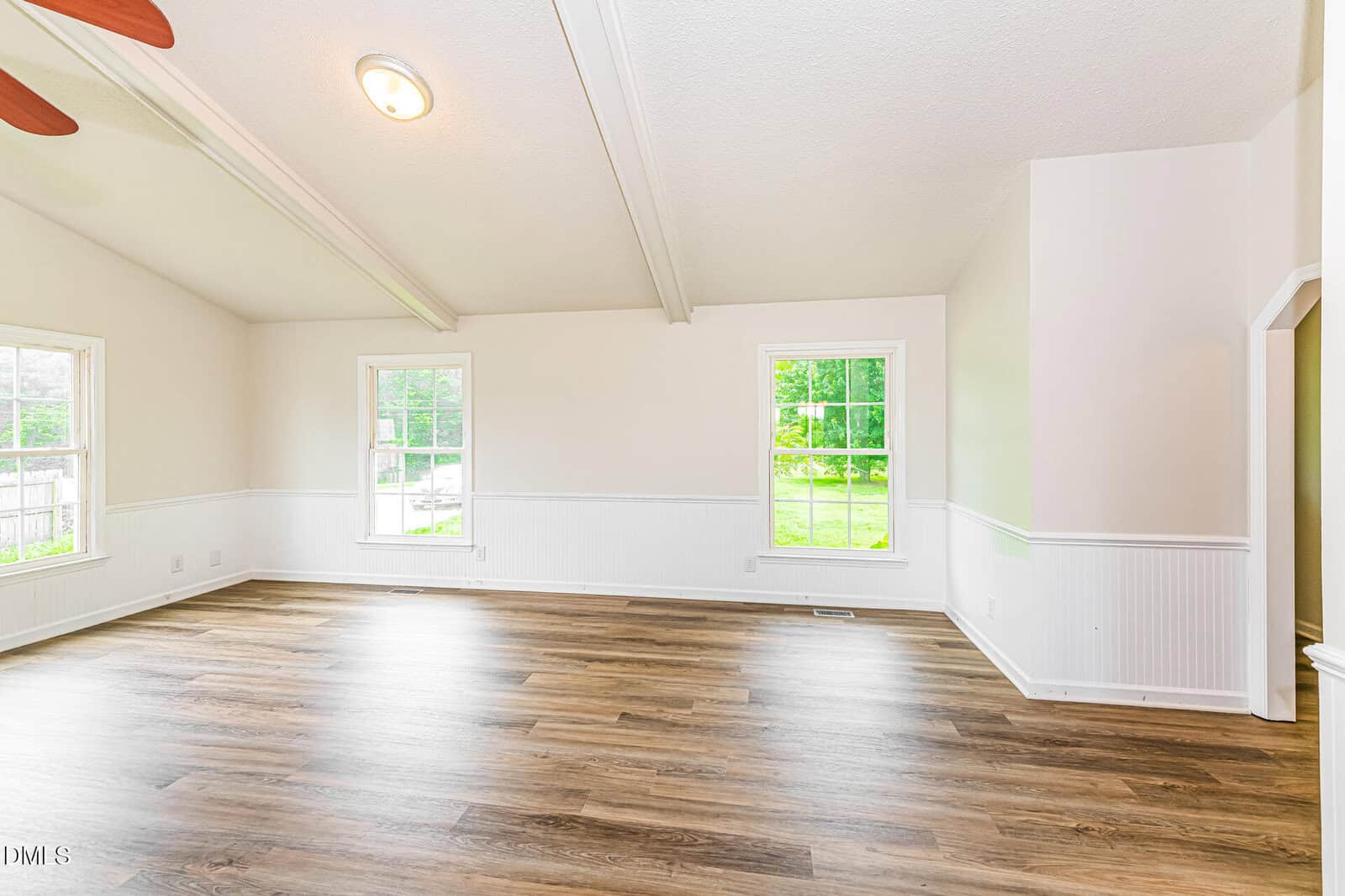 Bright, empty room with white walls, wood-style flooring, high ceiling with exposed beams, and three large windows showing green trees outside.