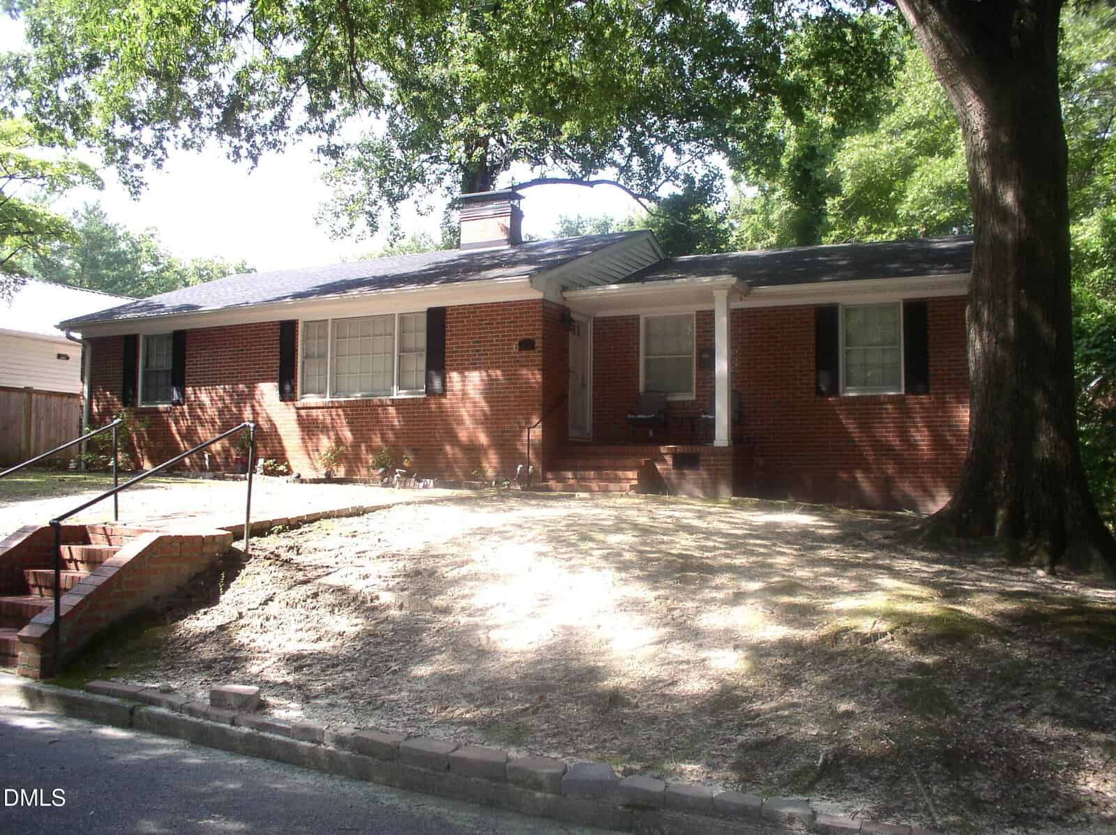 Charming single-story red brick house at 229 Perry Street in Henderson, featuring black shutters, a chimney, a covered front porch, steps with a metal railing, and large shaded trees in the front yard.
