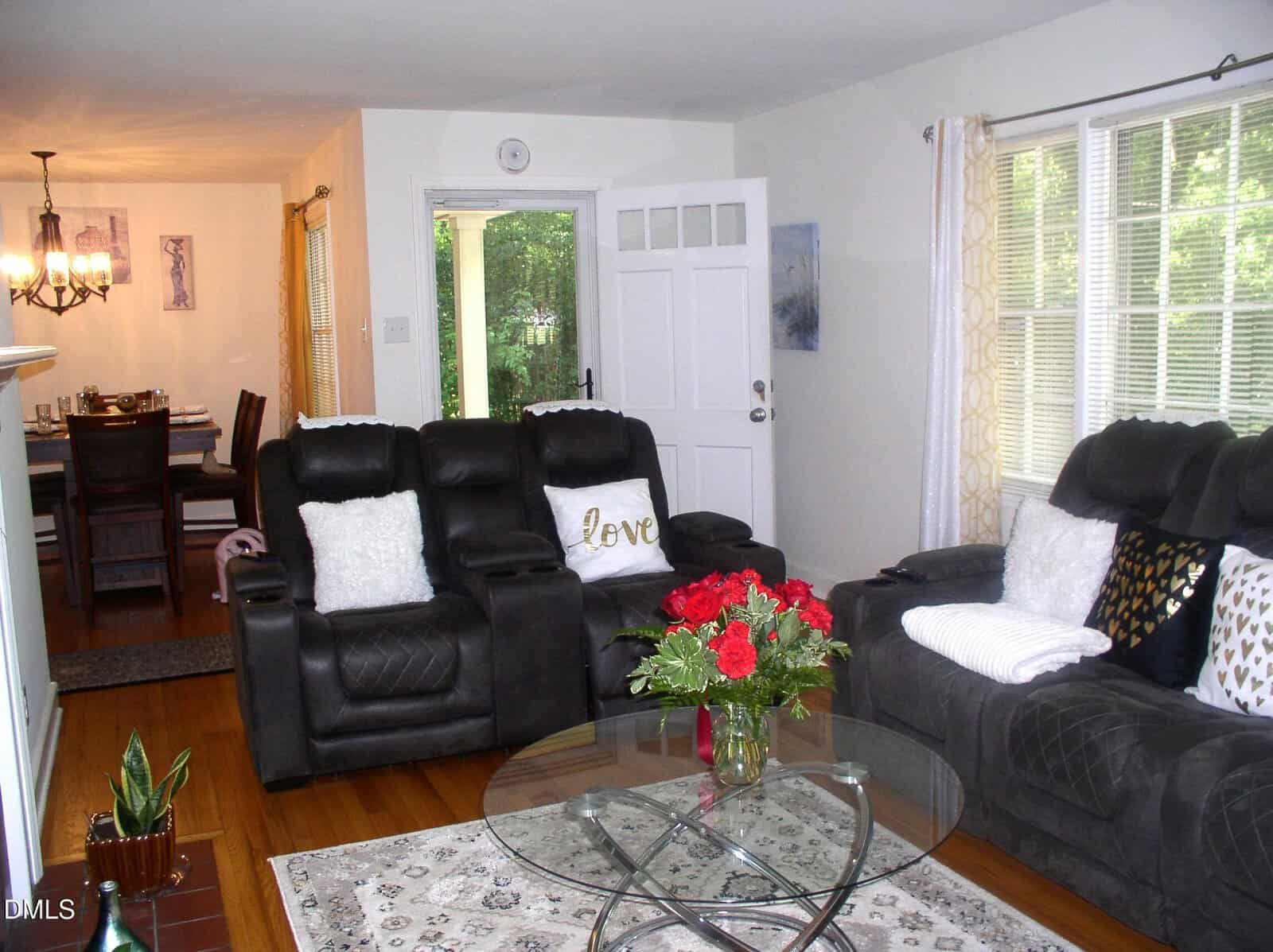 Living room with dark sofas, glass coffee table holding a vase of red roses, light rug, and large window. Dining area with table and chandelier is visible in the background. Door is open to outside greenery.