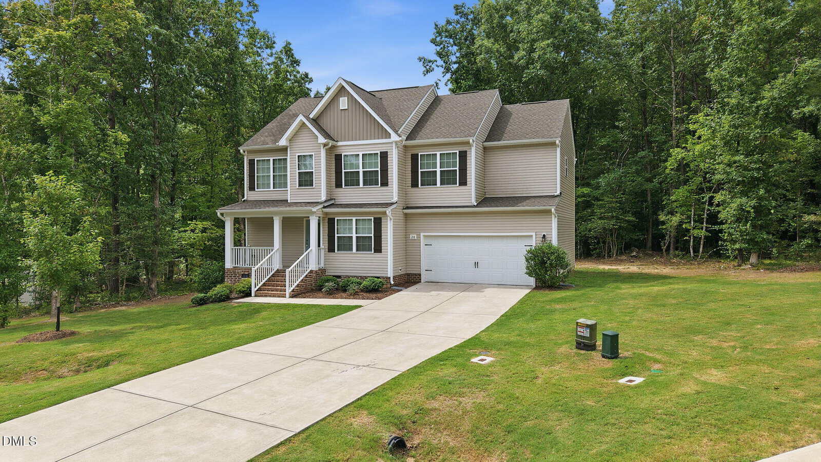 Two-story beige house with white trim, attached garage, and front porch. Concrete driveway curves through a well-maintained lawn, surrounded by tall green trees under a blue sky.