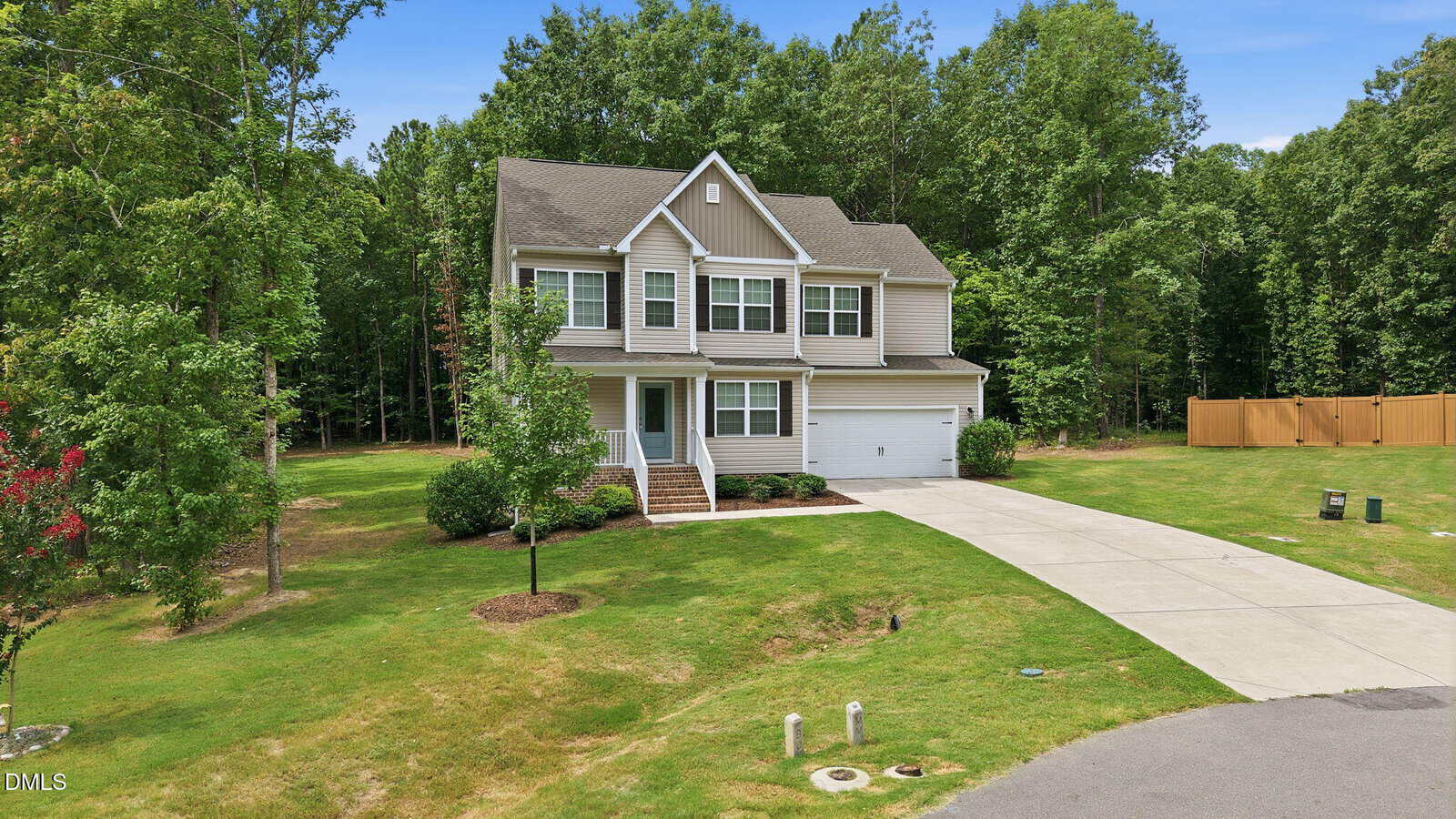 Two-story beige house with white trim, attached garage, and front porch, surrounded by a green lawn, trees, and a wooded area. Curved driveway leads from the street to the garage. Blue sky overhead.