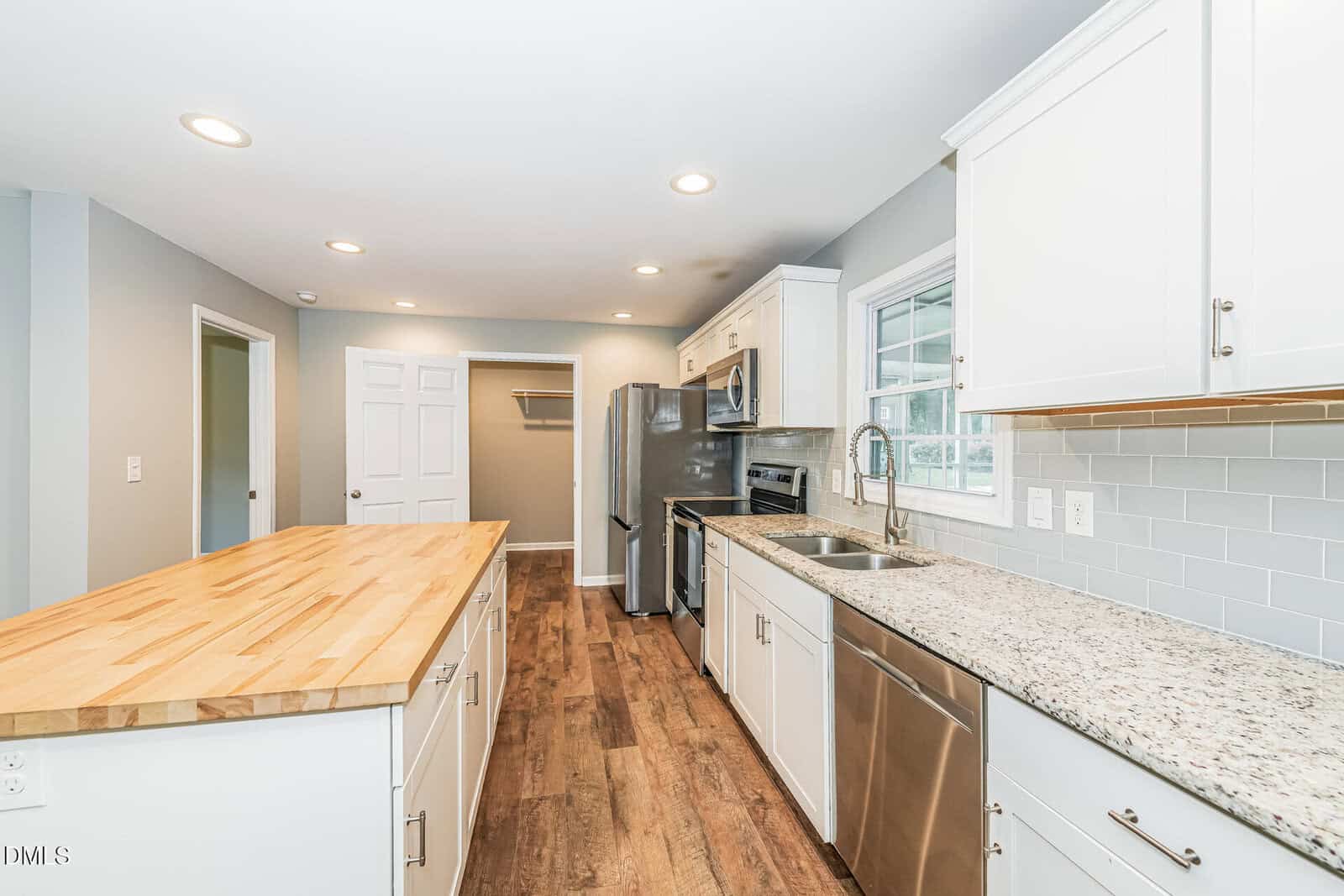 Modern kitchen with white cabinets, stainless steel appliances, a butcher block island, granite countertops, wood floors, and grey walls. A window is above the sink, and recessed ceiling lights brighten the space.