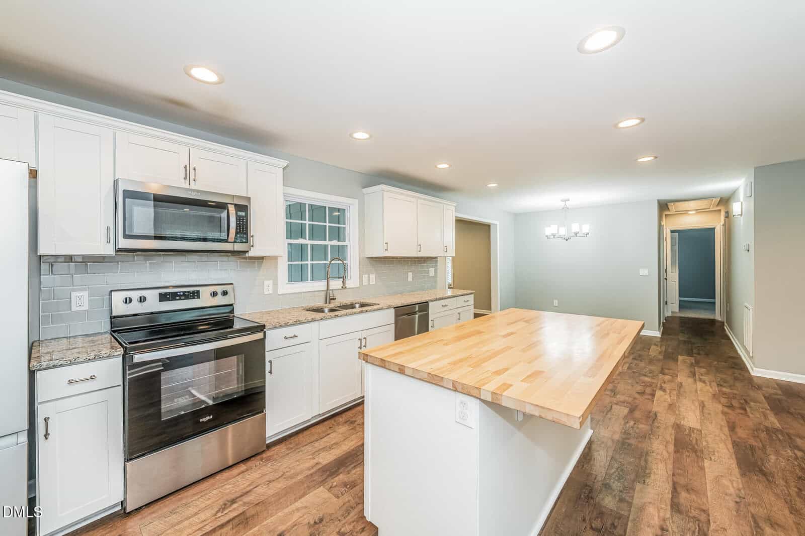 Modern kitchen with white cabinets, stainless steel appliances, a large wooden island, wood flooring, and recessed lighting. Dining area and hallway visible in the background.