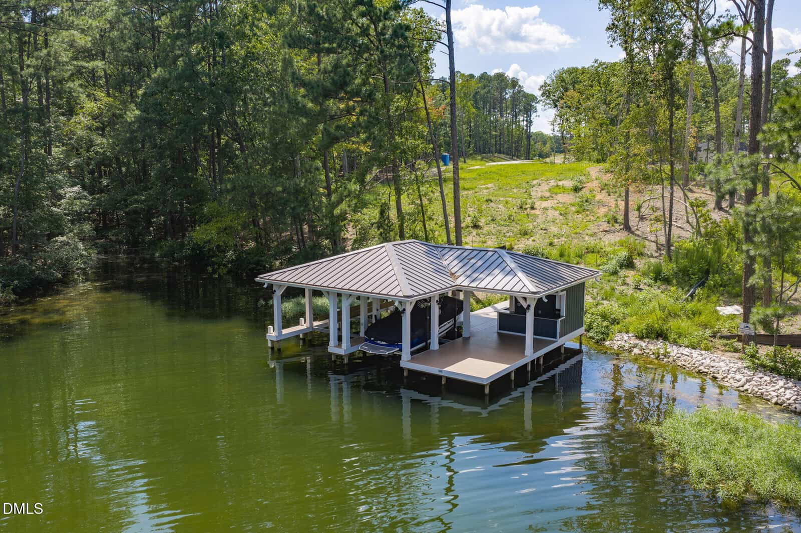 A covered boat dock with a metal roof extends over calm green water at 140 Serenity Point in Littleton, surrounded by trees and a grassy, slightly sloped shoreline under a partly cloudy sky.