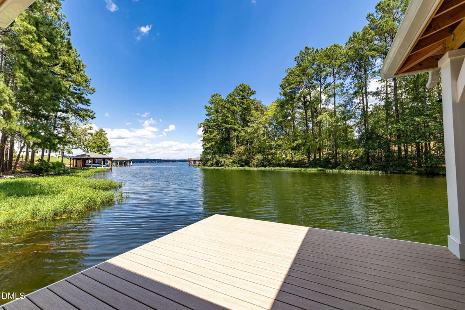 A wooden dock extends over a calm lake, surrounded by lush green trees under a bright blue sky with scattered white clouds.
