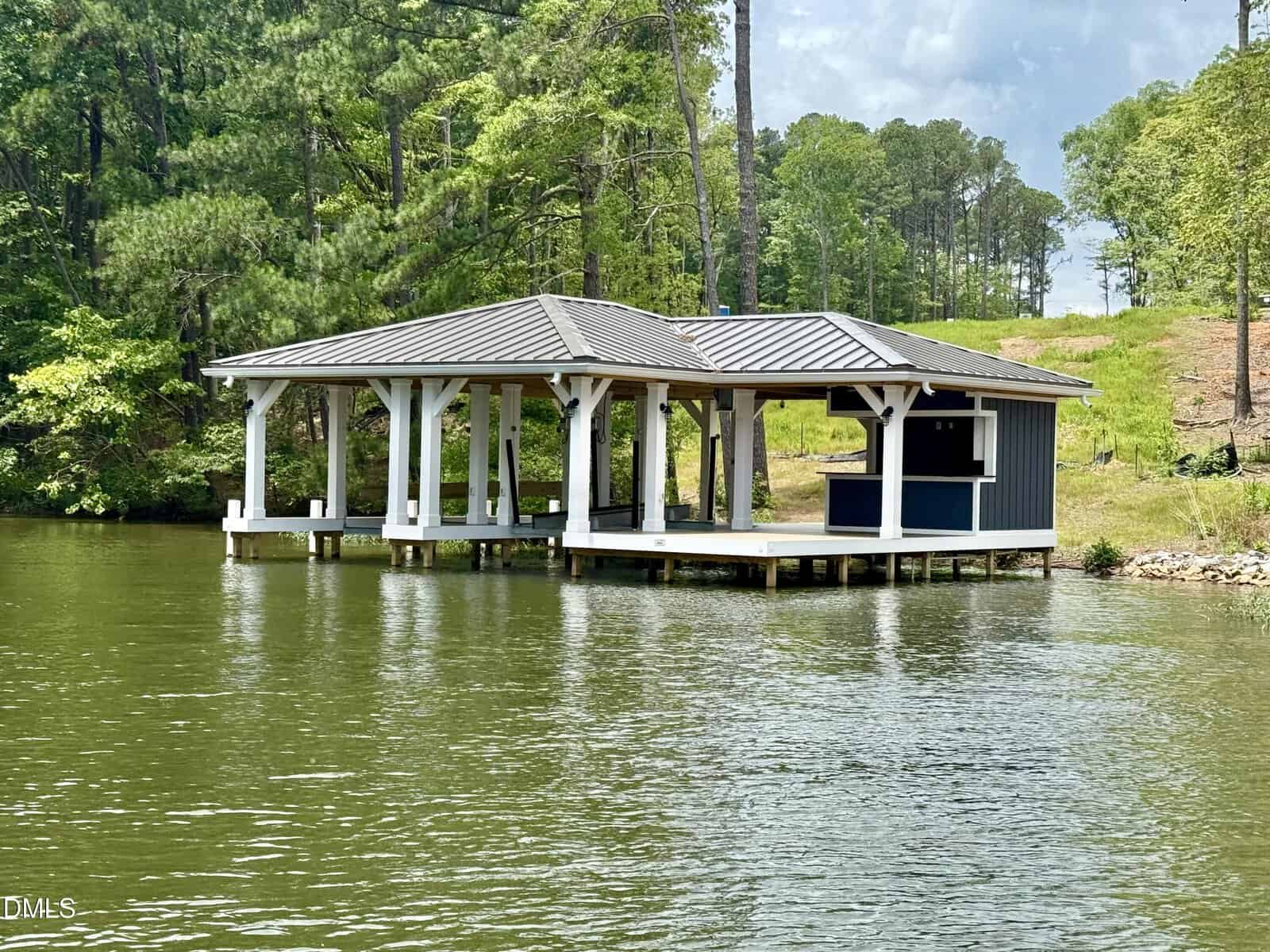 A lakeside covered dock with a boat slip and small enclosed section, surrounded by trees and greenery, with calm water in the foreground.