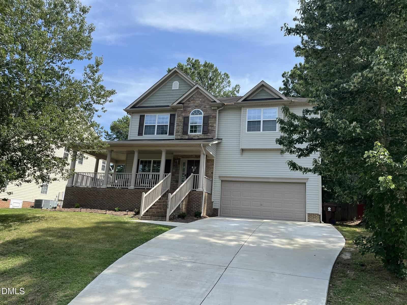 Two-story suburban house at 1708 Marlow Road in Creedmoor, featuring beige siding, brick accents, a covered front porch, and a double garage. A concrete driveway curves up to the entrance, surrounded by trees and a grassy yard under a blue sky.