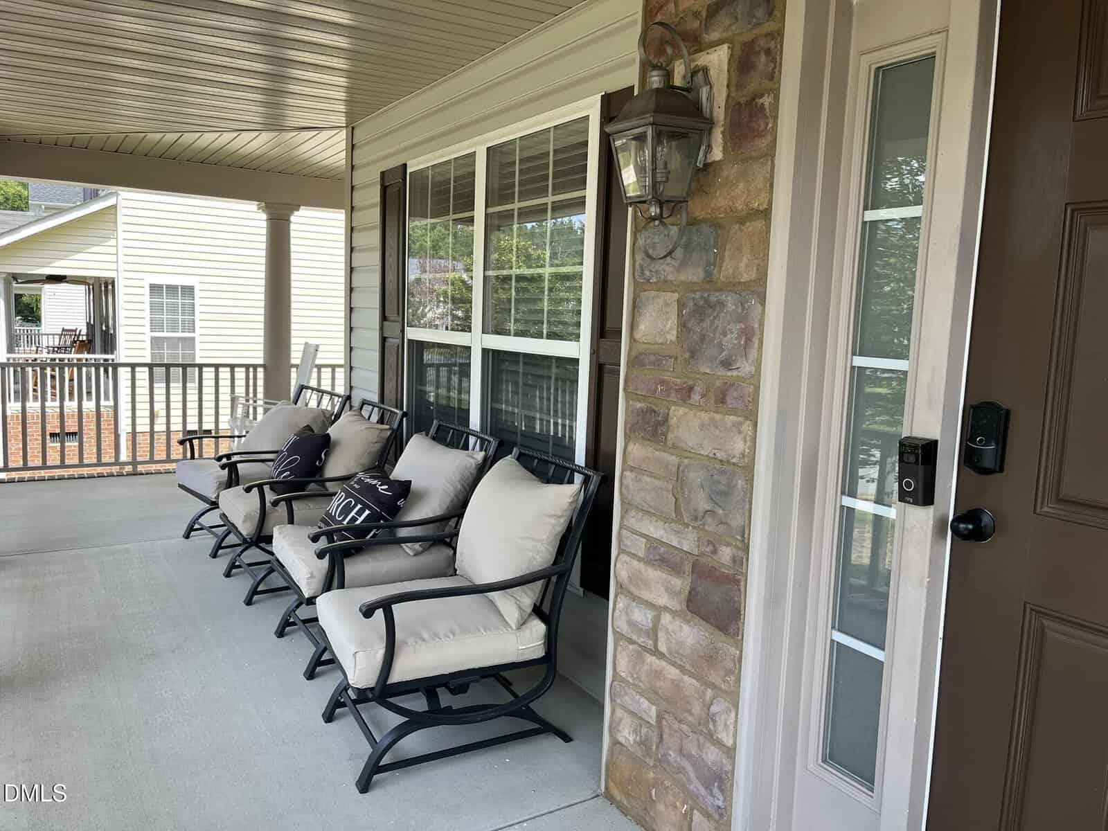 A covered porch with four cushioned rocking chairs, each with a pillow, sits beside a stone wall and a front door. Large windows and a lantern-style light fixture are visible.