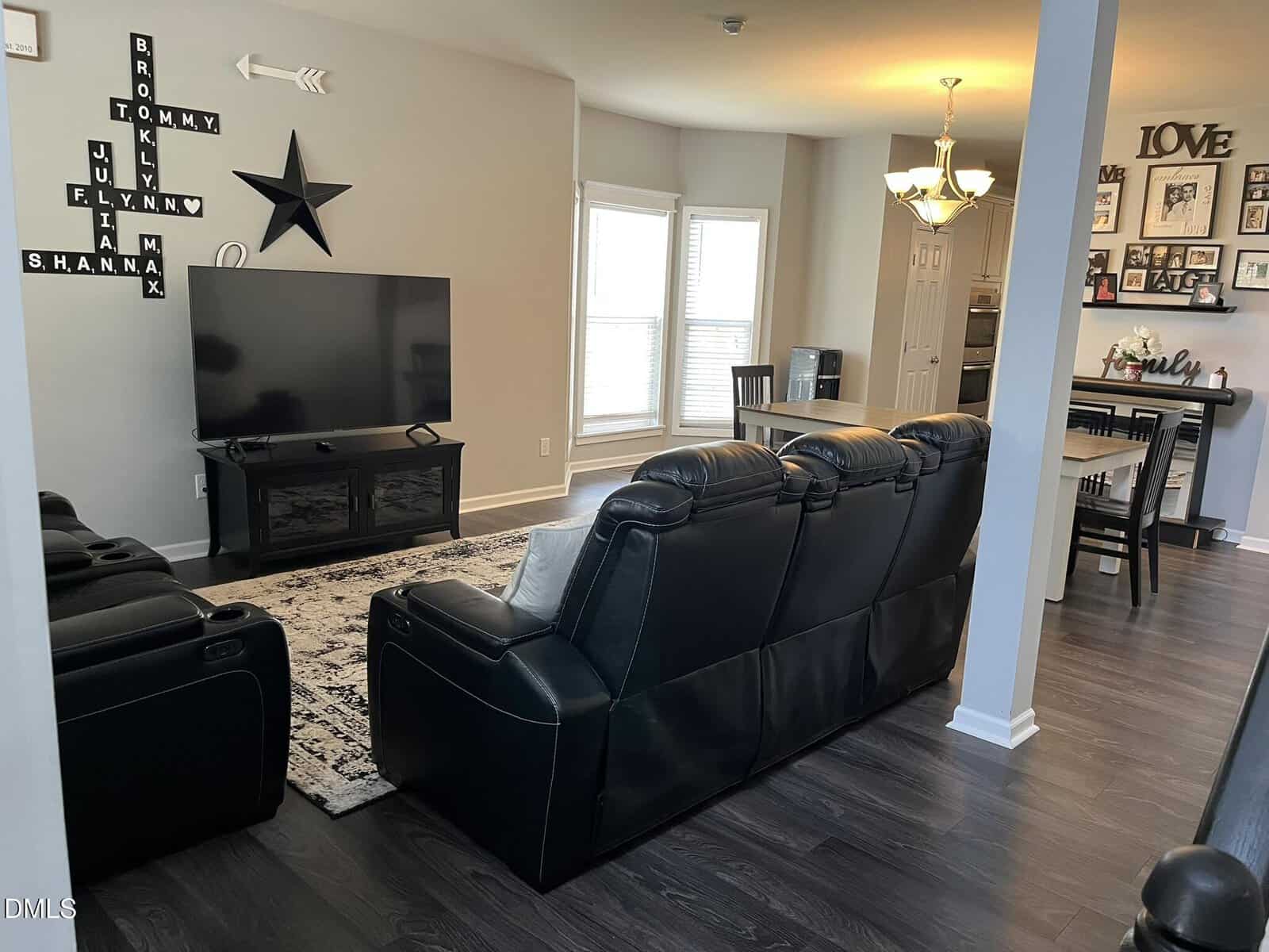 Modern living room with black leather sofas facing a large TV, wall decor with names, a star, and “LOVE” art. Dining area with table and chairs is visible, along with a kitchen in the background.