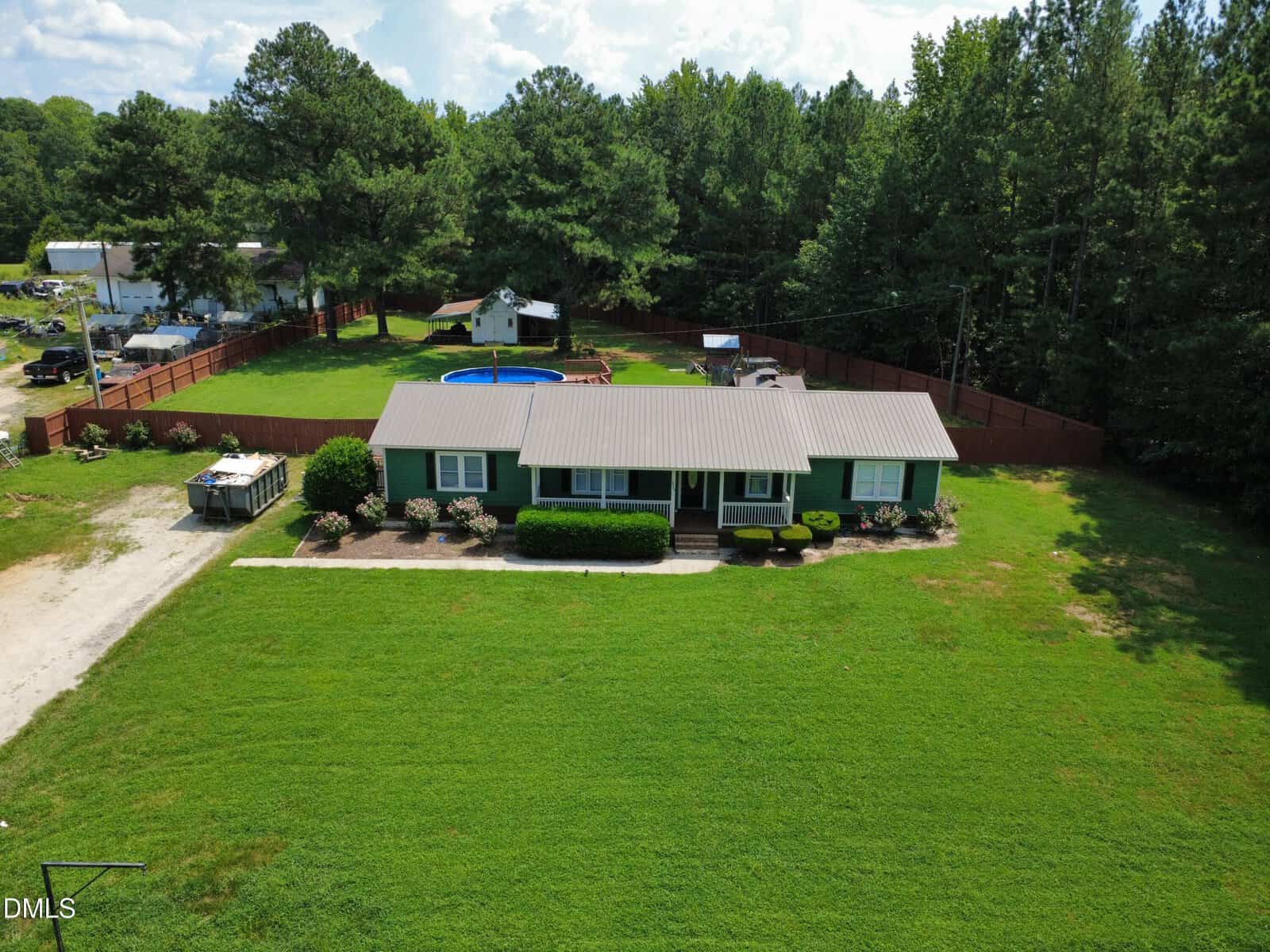 A green single-story house with a gray roof sits on a large lawn, bordered by a brown fence. There is a blue above-ground pool and a shed in the fenced backyard, surrounded by tall trees.