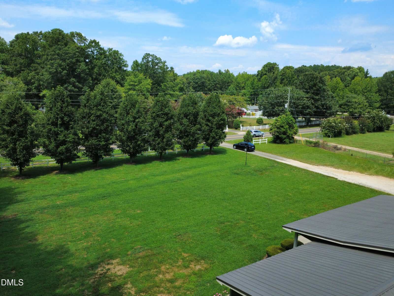 A green yard with a row of trees along a road, a black car on a gravel driveway, and distant trees under a partly cloudy sky.