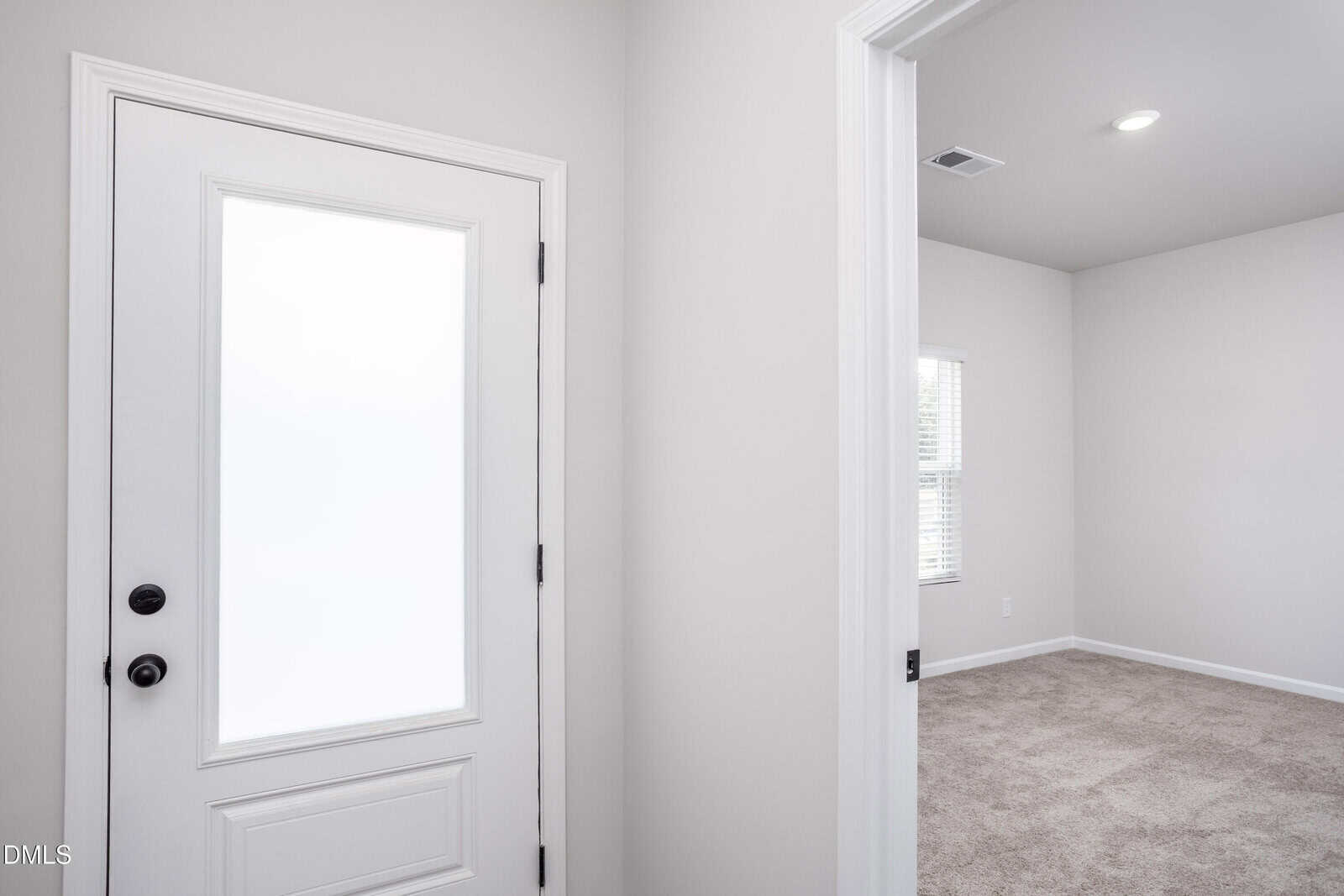 A white front door with a frosted glass panel opens into a bright room with light-colored walls, white trim, beige carpet, and a window with blinds letting in natural light.