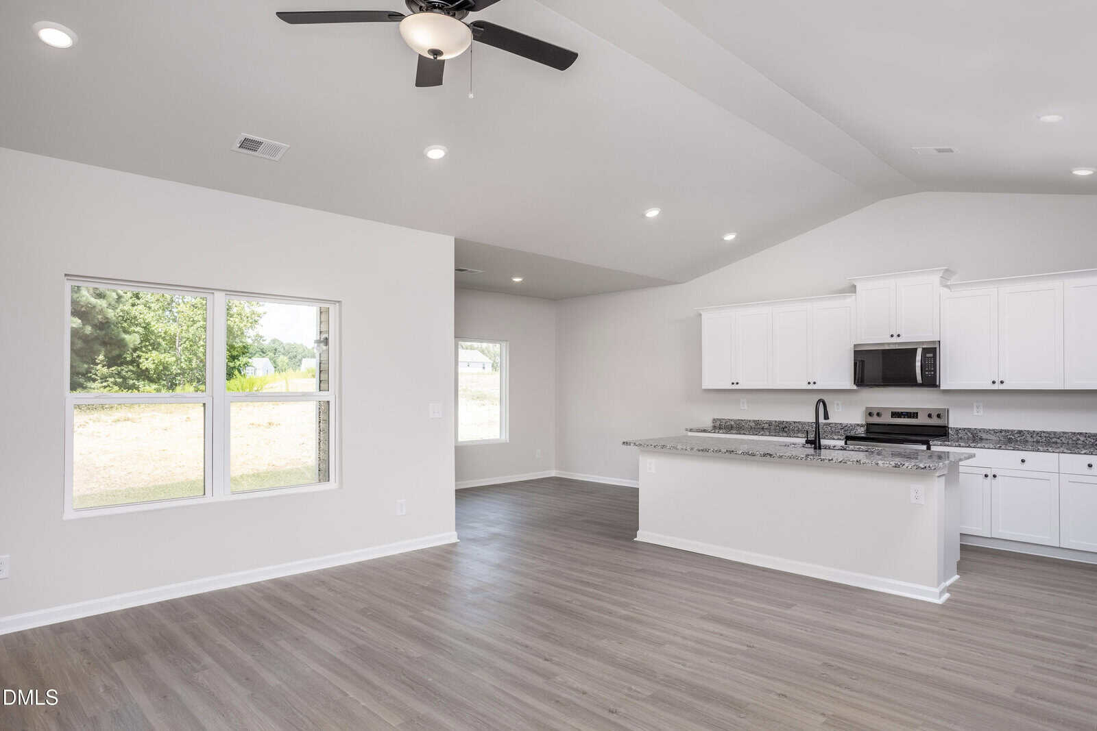 Modern, empty kitchen and living area with white cabinets, stainless steel appliances, granite countertops, wood flooring, ceiling fan, and large windows letting in natural light.