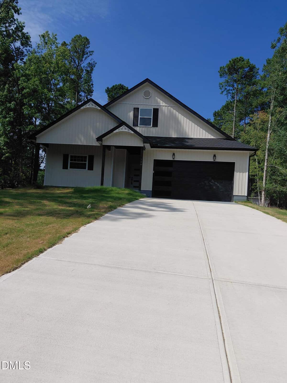 Modern single-story house with black and white exterior, a dark garage door, and a steep driveway. Surrounded by green grass and tall trees under a clear blue sky.