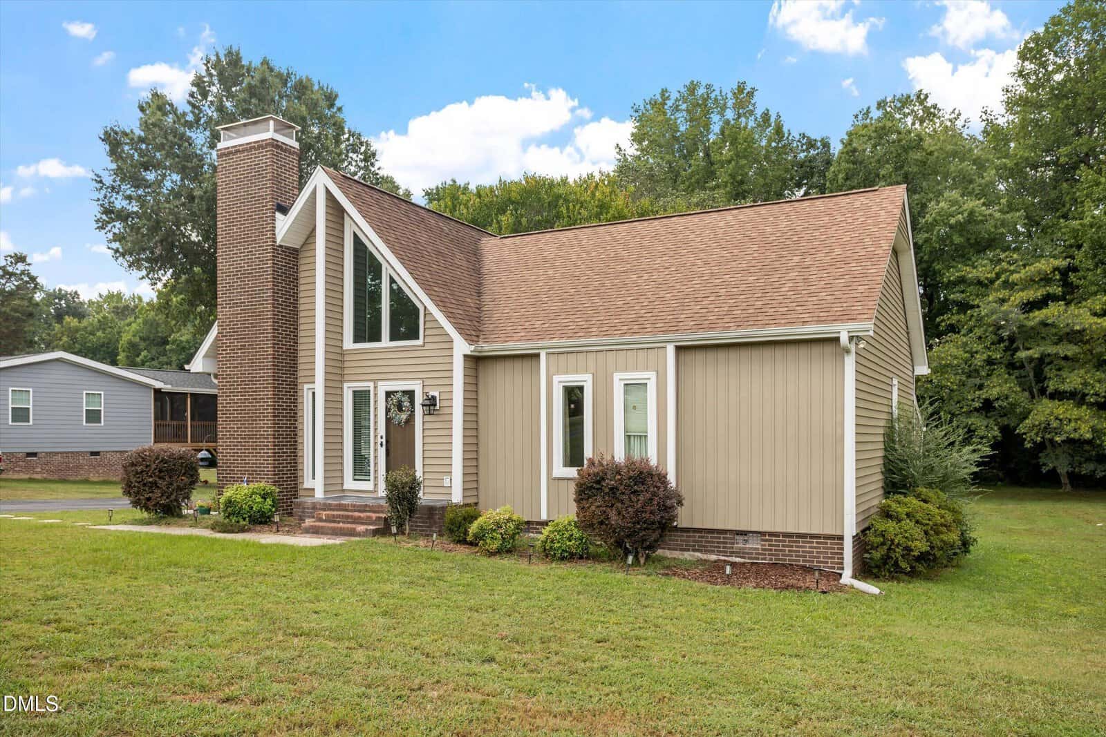 Modern tan house with a steep roof, large front windows, and a brick chimney, surrounded by green lawn and bushes, with trees and another house in the background under a partly cloudy sky.