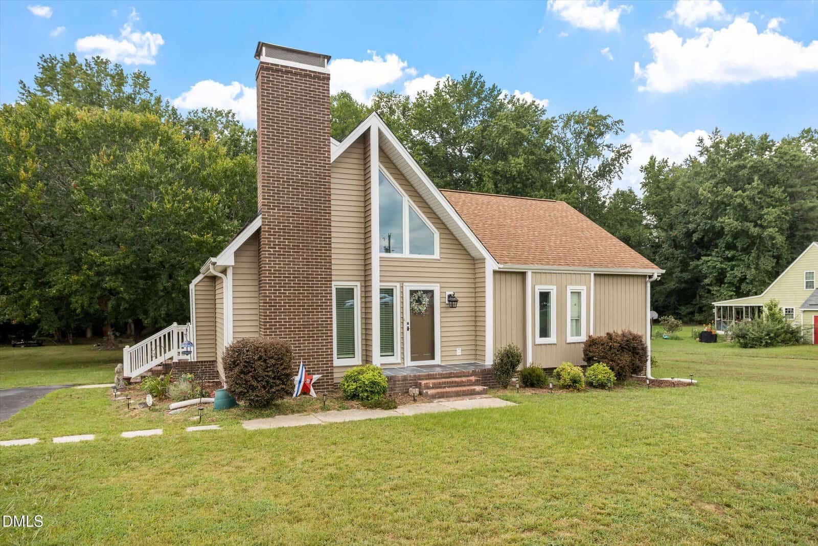 Modern beige house with large windows, a tall brick chimney, triangular roof, and a well-kept lawn. Steps and small shrubs lead to the front door. Trees and another house are visible in the background under a blue sky.