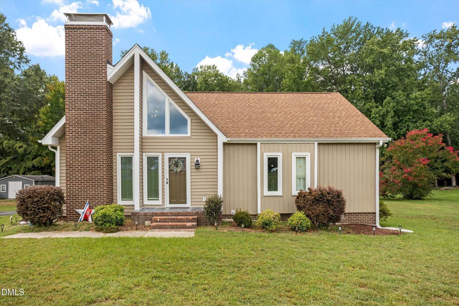 A tan house with a steep roof, large front windows, and a brick chimney sits on a grassy lawn at 116 Bridle Trail in Youngsville, surrounded by shrubs and trees in the background.