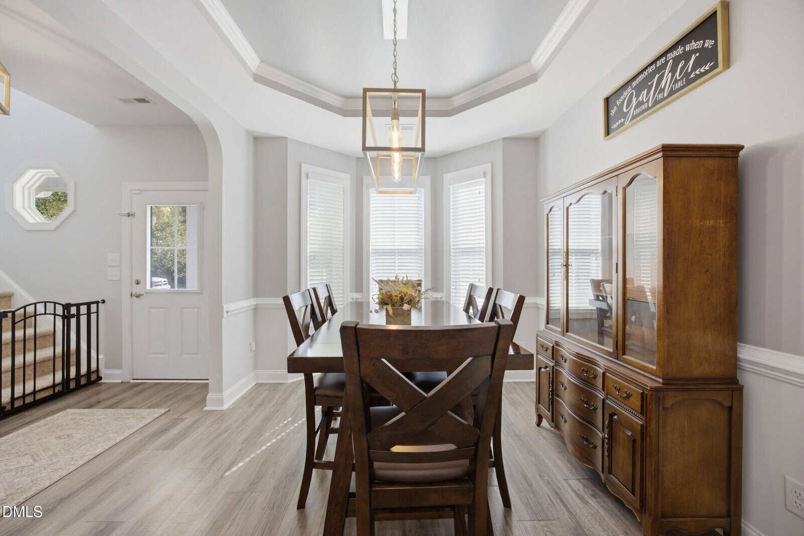 Bright dining room with wooden table and chairs, a china cabinet, decorative light fixture, bay windows with blinds, and light gray walls with white trim. A black safety gate is near the entrance door.