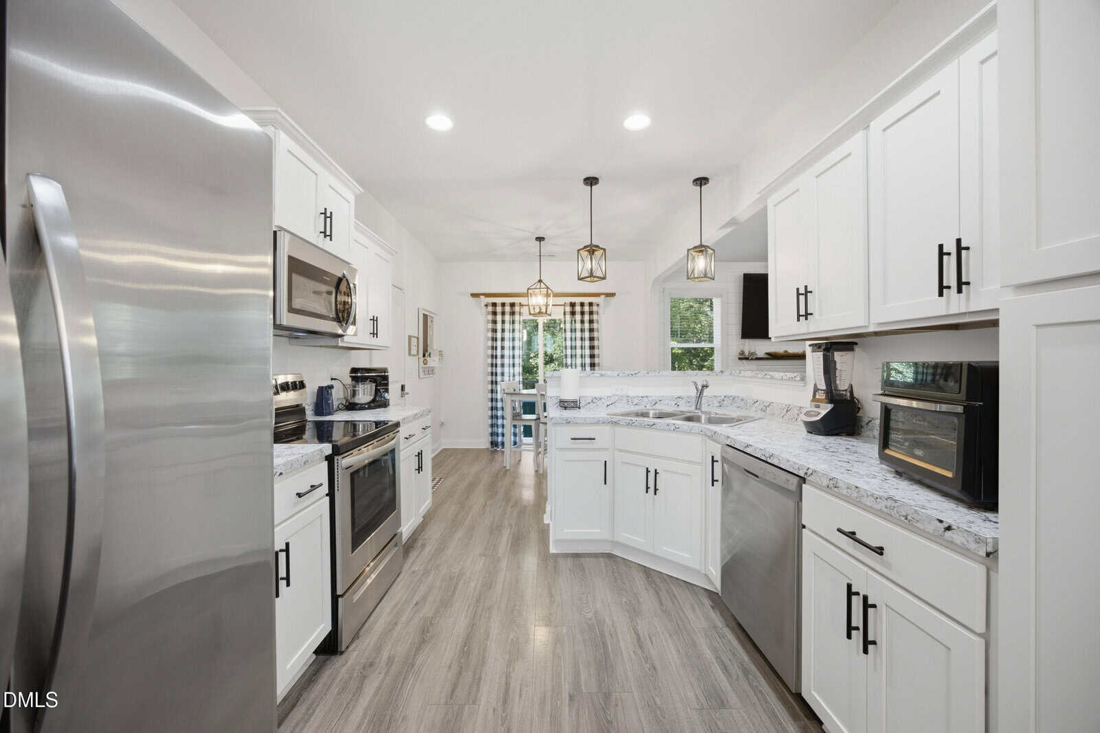 Modern white kitchen with stainless steel appliances, marble countertops, pendant lights, and wood-style flooring. A window and glass door provide natural light at the far end of the room.