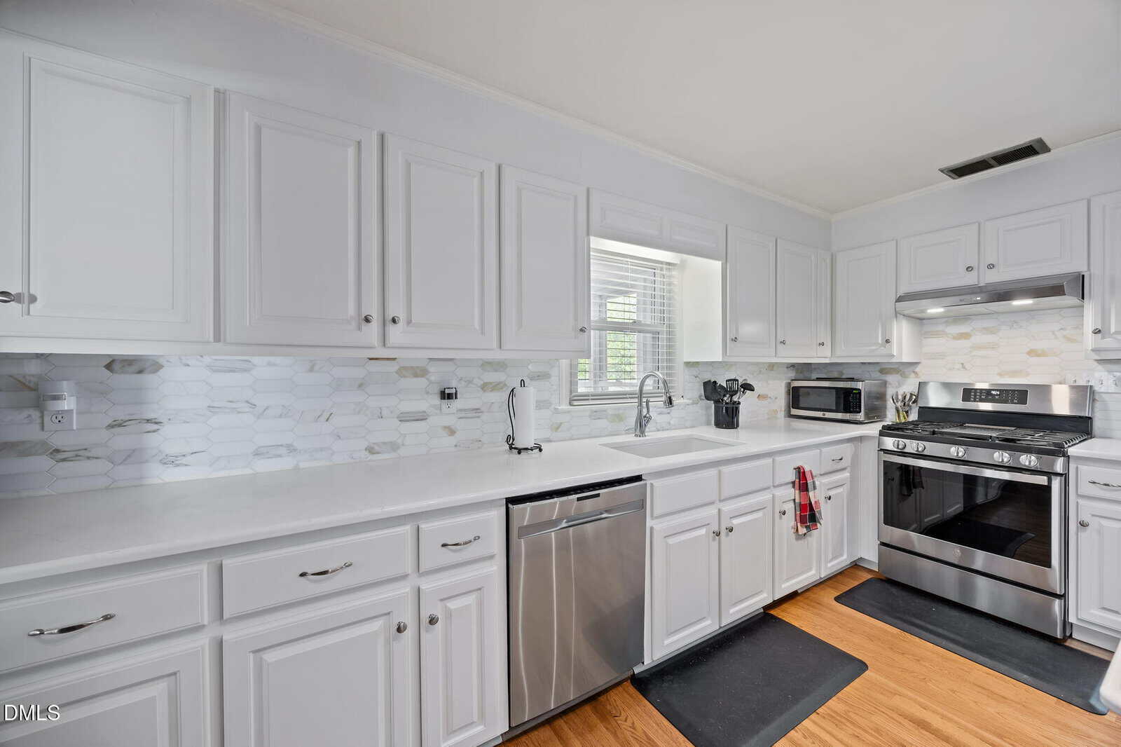 Modern kitchen with white cabinets, stainless steel appliances, tile backsplash, sink under a window, and light wood flooring. A red towel hangs on a drawer handle near the dishwasher.
