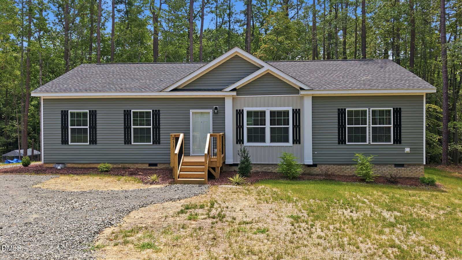 A single-story gray house with black shutters, a front porch with stairs, and surrounded by trees. There is a gravel driveway and patchy grass in the front yard.
