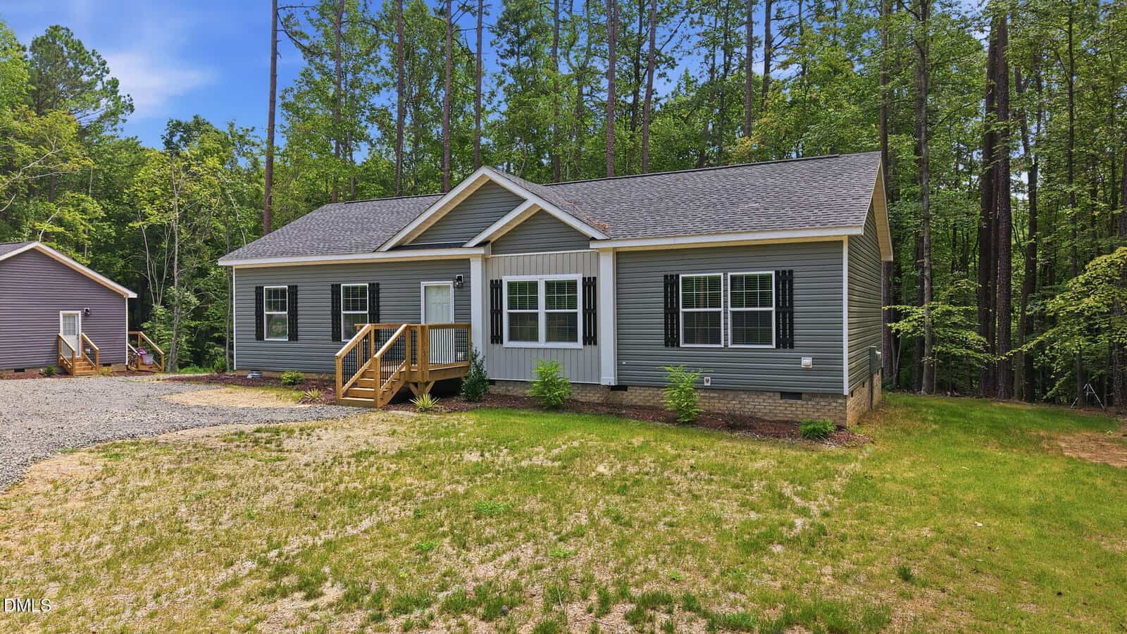 Single-story gray house with white trim, black shutters, and a small wooden porch, surrounded by tall trees and greenery. Gravel driveway and patchy grass are visible in the front yard.