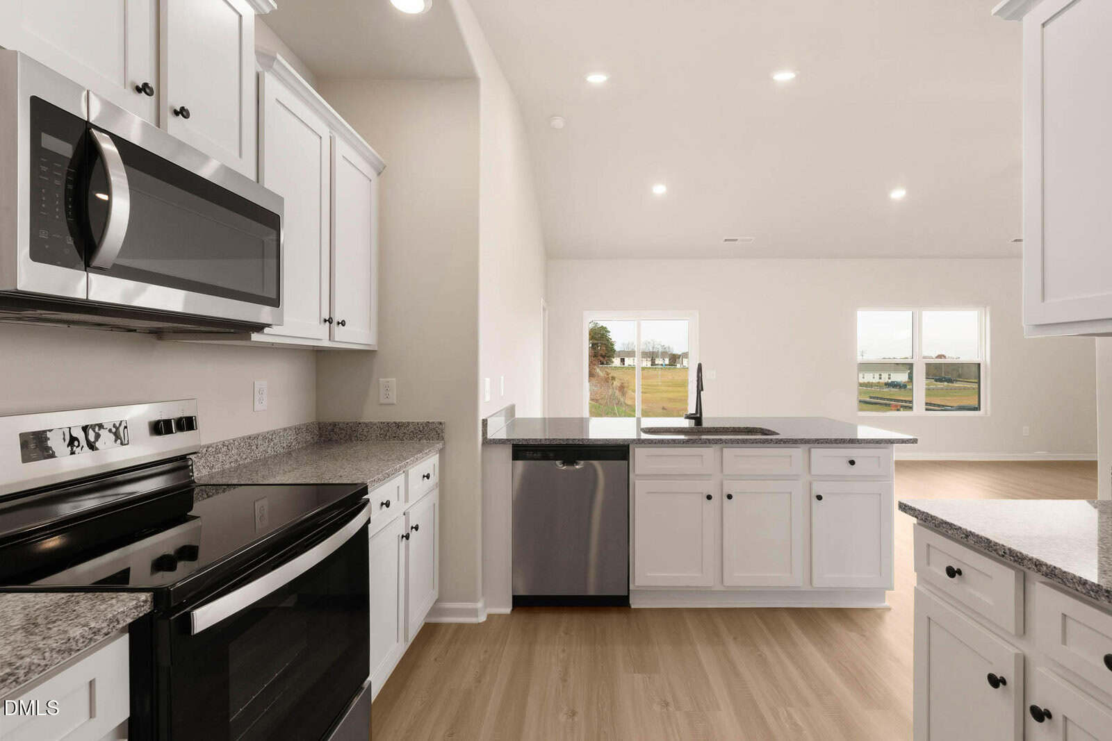Modern kitchen with white cabinets, stainless steel appliances, granite countertops, a center island with sink and dishwasher, light wood flooring, and large windows letting in natural light, facing an open living area.