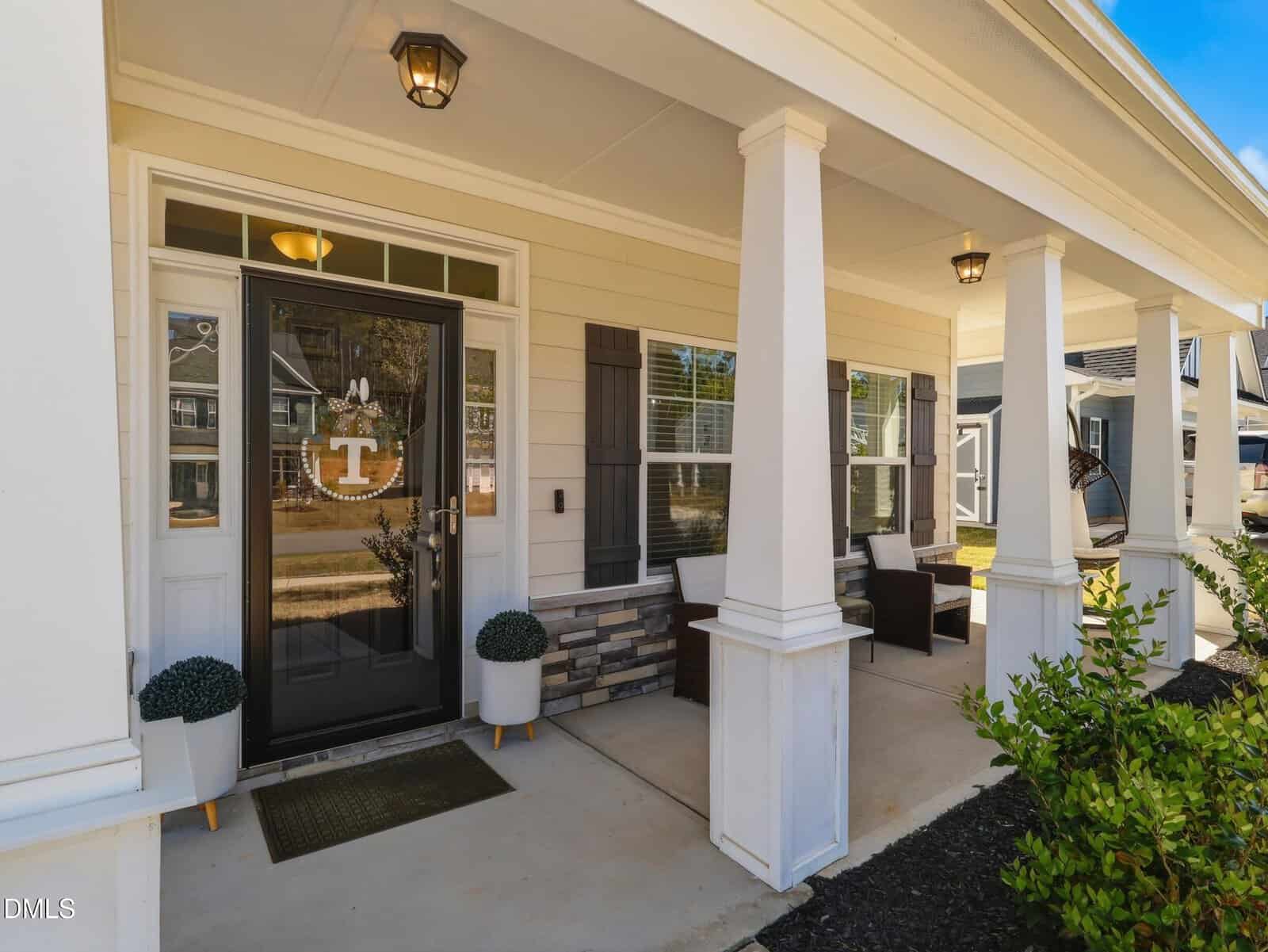 Front porch of a house with white columns, a black front door with a wreath, potted plants, outdoor seating, windows with shutters, and a porch light. Other houses and trees are visible in the background.