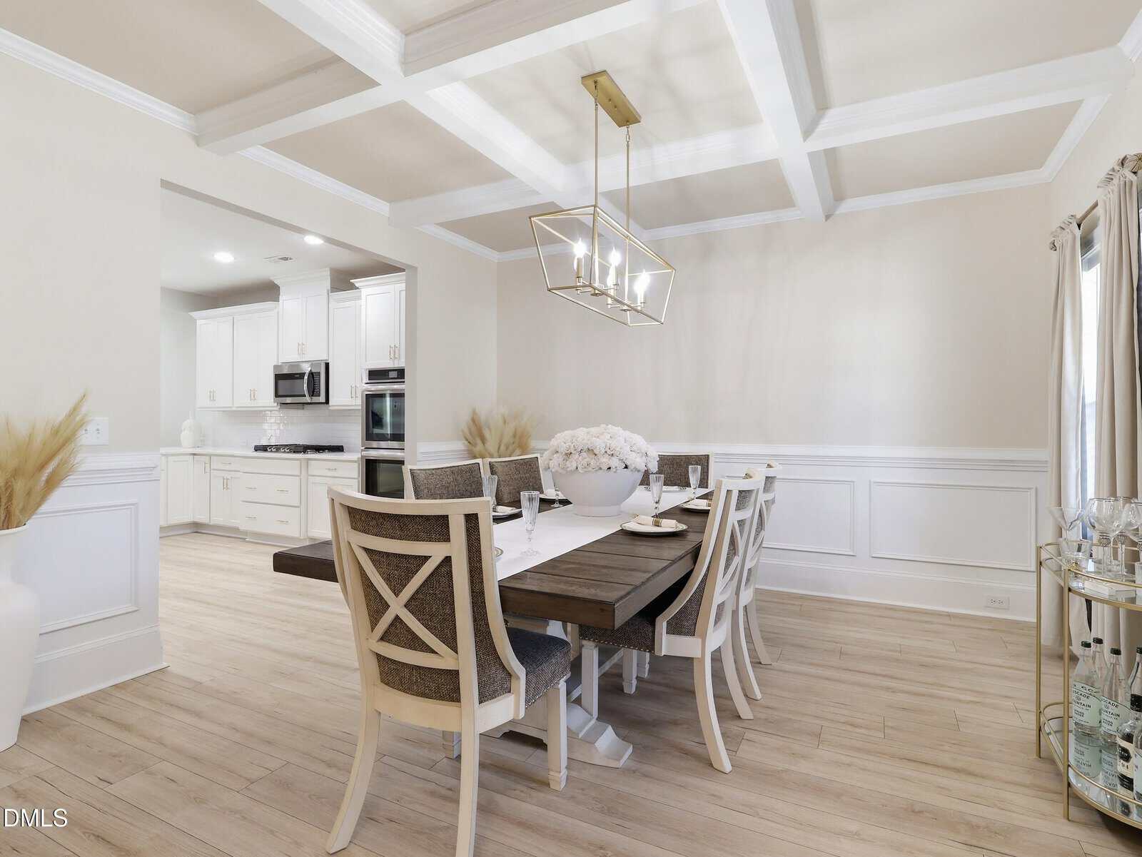 A modern dining room with a dark wood table, eight chairs, a large white floral centerpiece, white paneled walls, light wood floors, and a gold geometric chandelier, connected to a white kitchen.