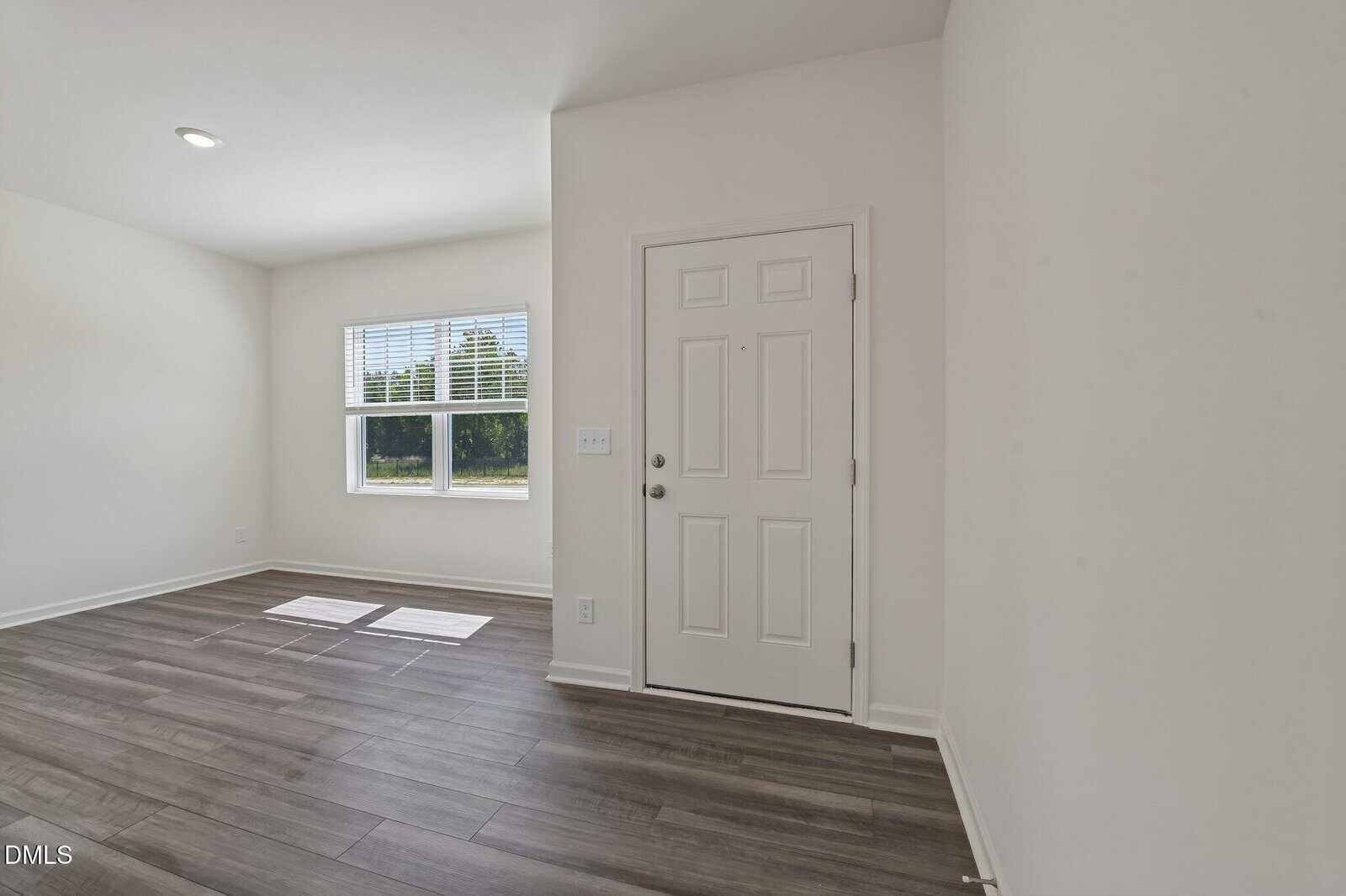 A bright, empty room with white walls, wood-style flooring, a window letting in sunlight, and a closed white front door.