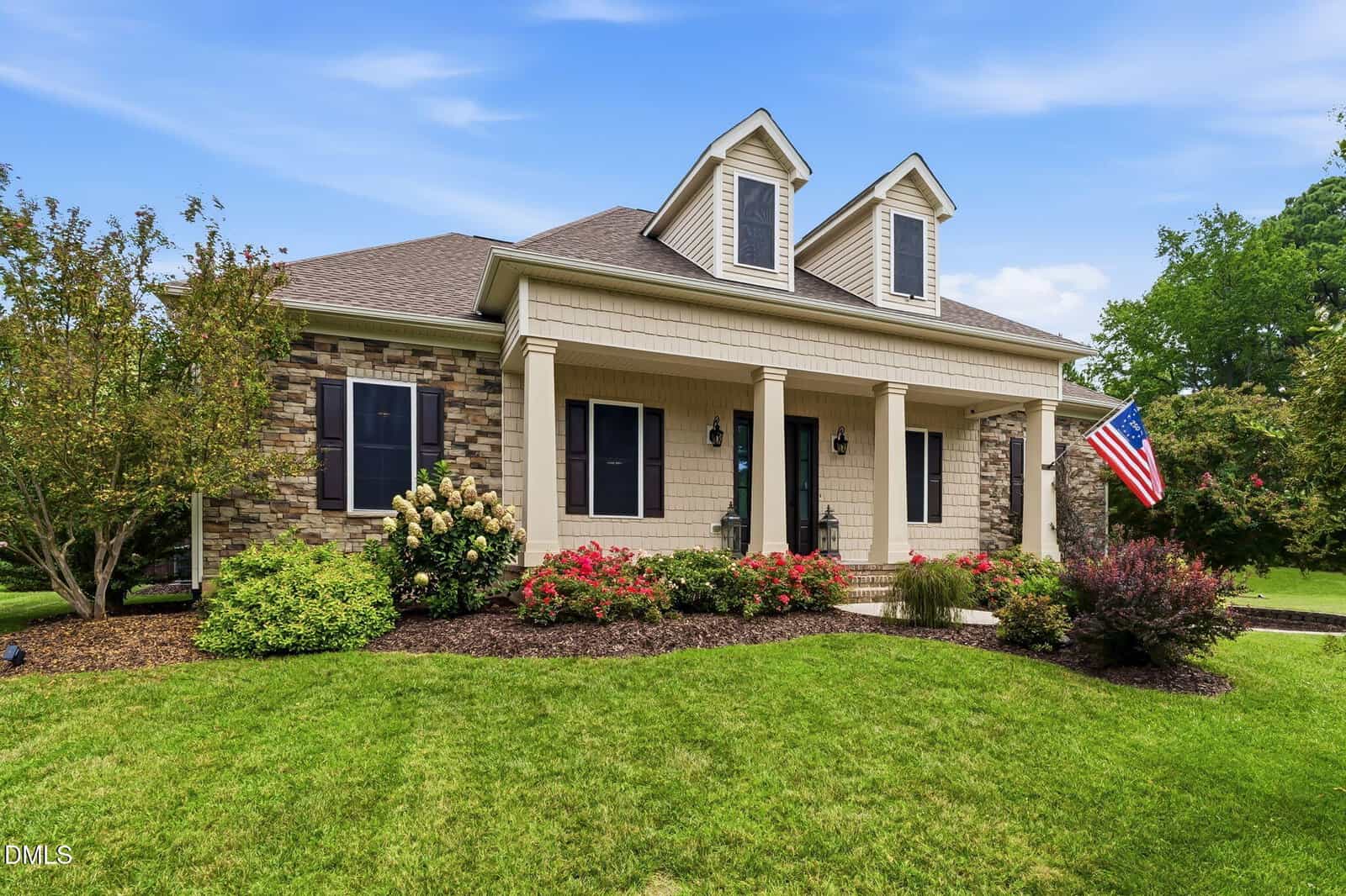 Single-story house with stone and beige siding, two dormer windows, a columned front porch, landscaped yard with bushes and flowers, and an American flag displayed near the entrance under a partly cloudy blue sky.