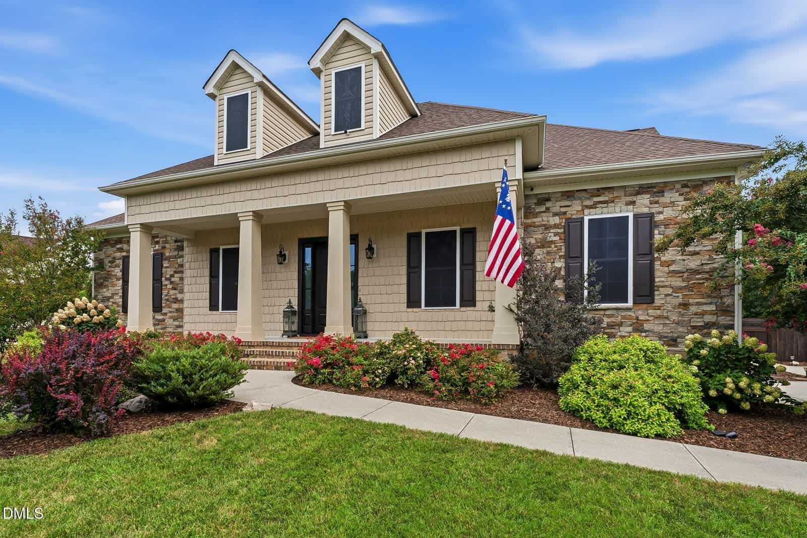 Single-story house with beige siding, stone accents, and two dormer windows. A front porch with columns, American flag, and landscaped yard with colorful bushes and green grass are visible under a blue sky at 4517 Carson Layne in Franklinton.