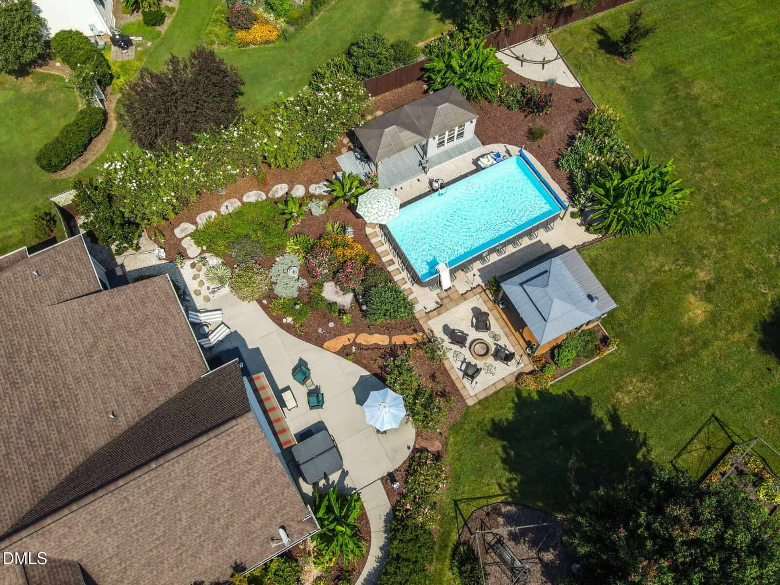 Aerial view of a landscaped backyard with a swimming pool, patio seating, gazebo, fire pit, garden beds, and lush green lawn surrounded by trees and shrubs.