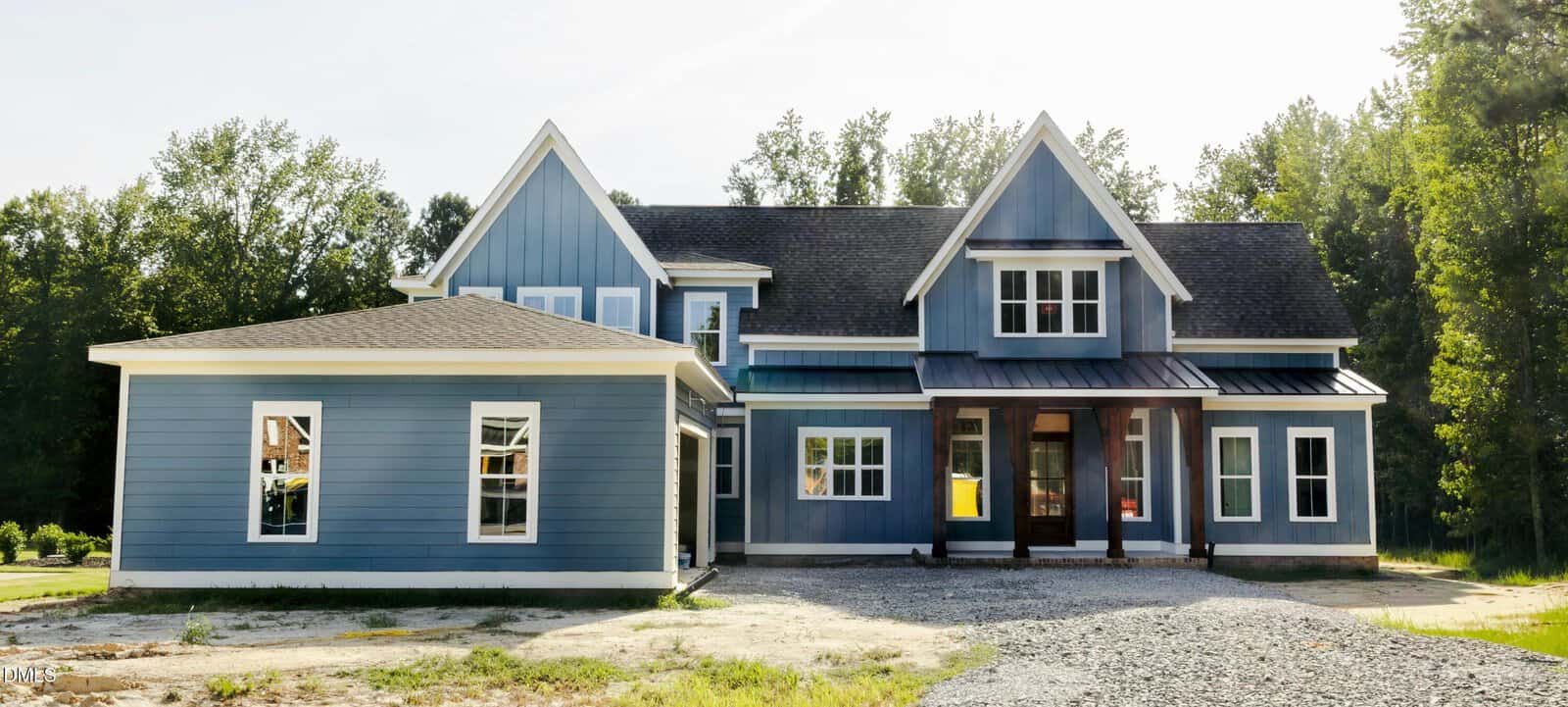 A two-story blue house with white trim sits on a gravel driveway, surrounded by trees and grass, with sunlight illuminating the front exterior.
