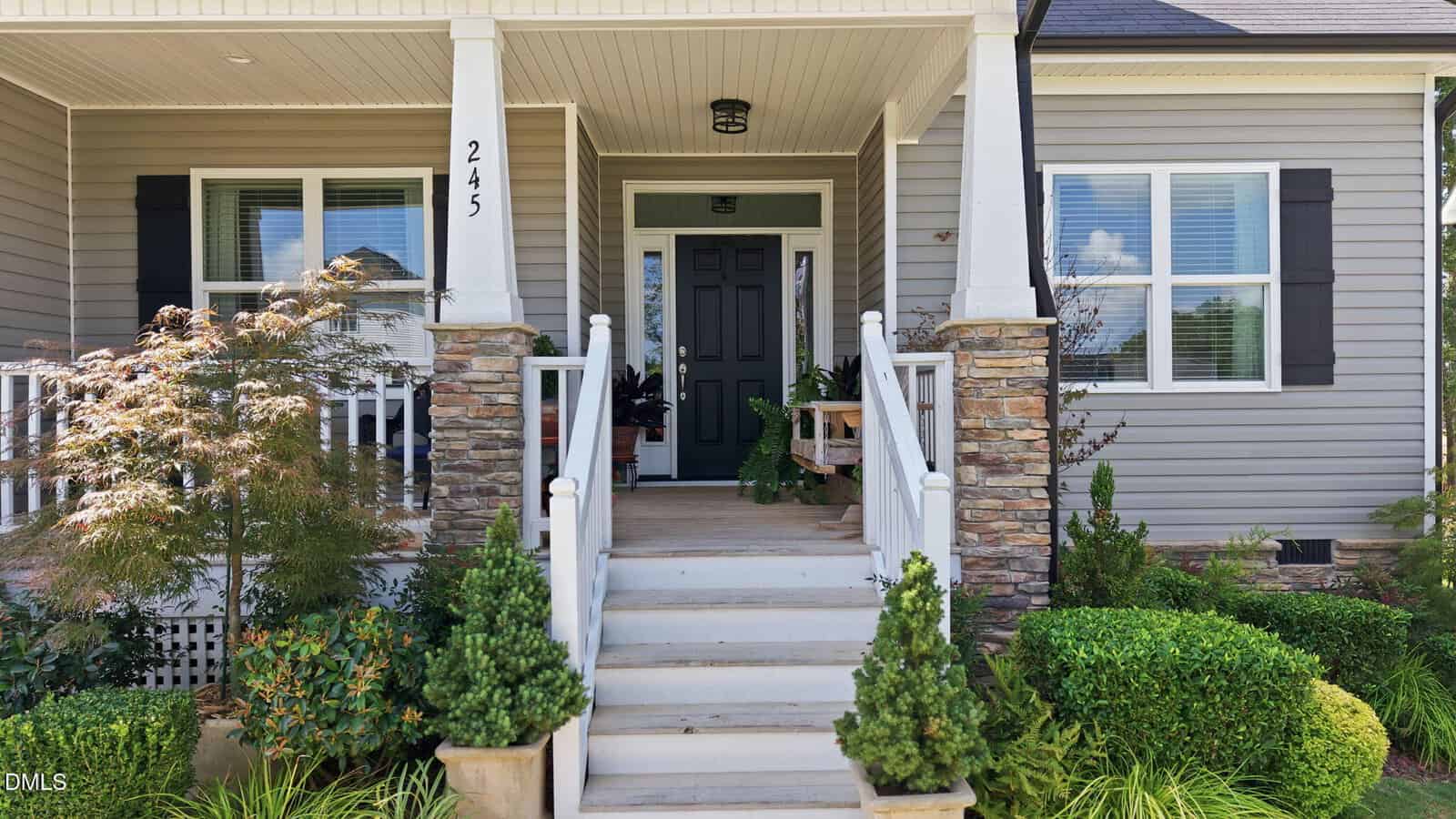 Front porch of a house with stone columns, white railings, green shrubs, small trees, and a black front door. House has gray siding and black shutters, with a house number “245” on a column.