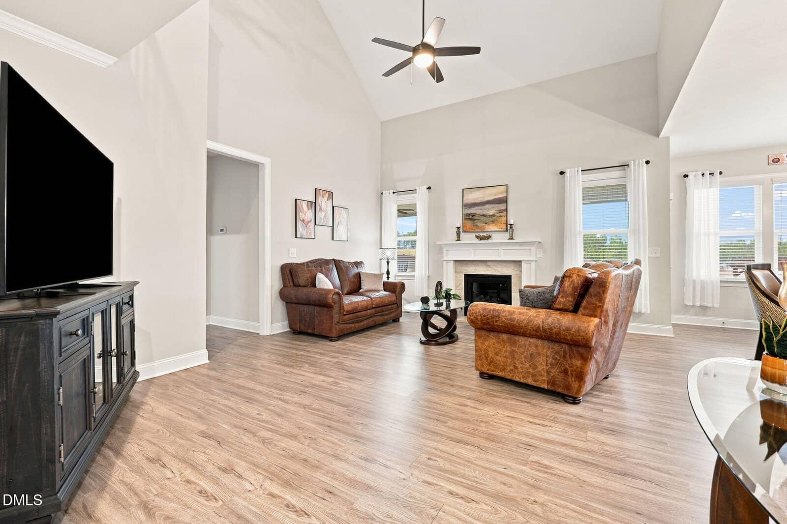 Spacious living room with brown leather furniture, large TV, fireplace, two windows with white curtains, wood flooring, ceiling fan, and neutral decor.