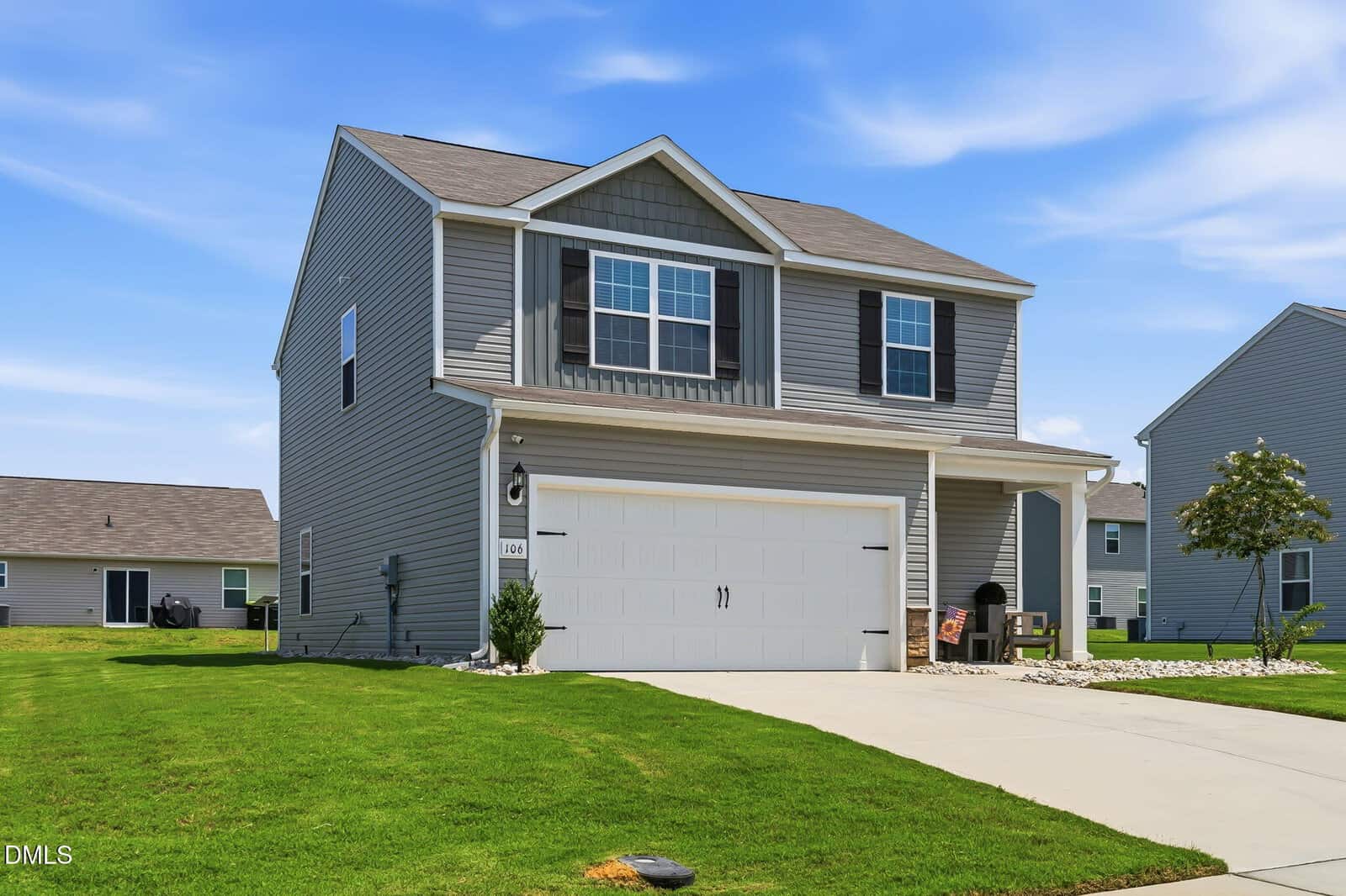 Two-story gray house with white trim, a double garage, and a covered front porch. Well-kept lawn, small shrubs by the entrance, and a bright blue sky with scattered clouds in the background.