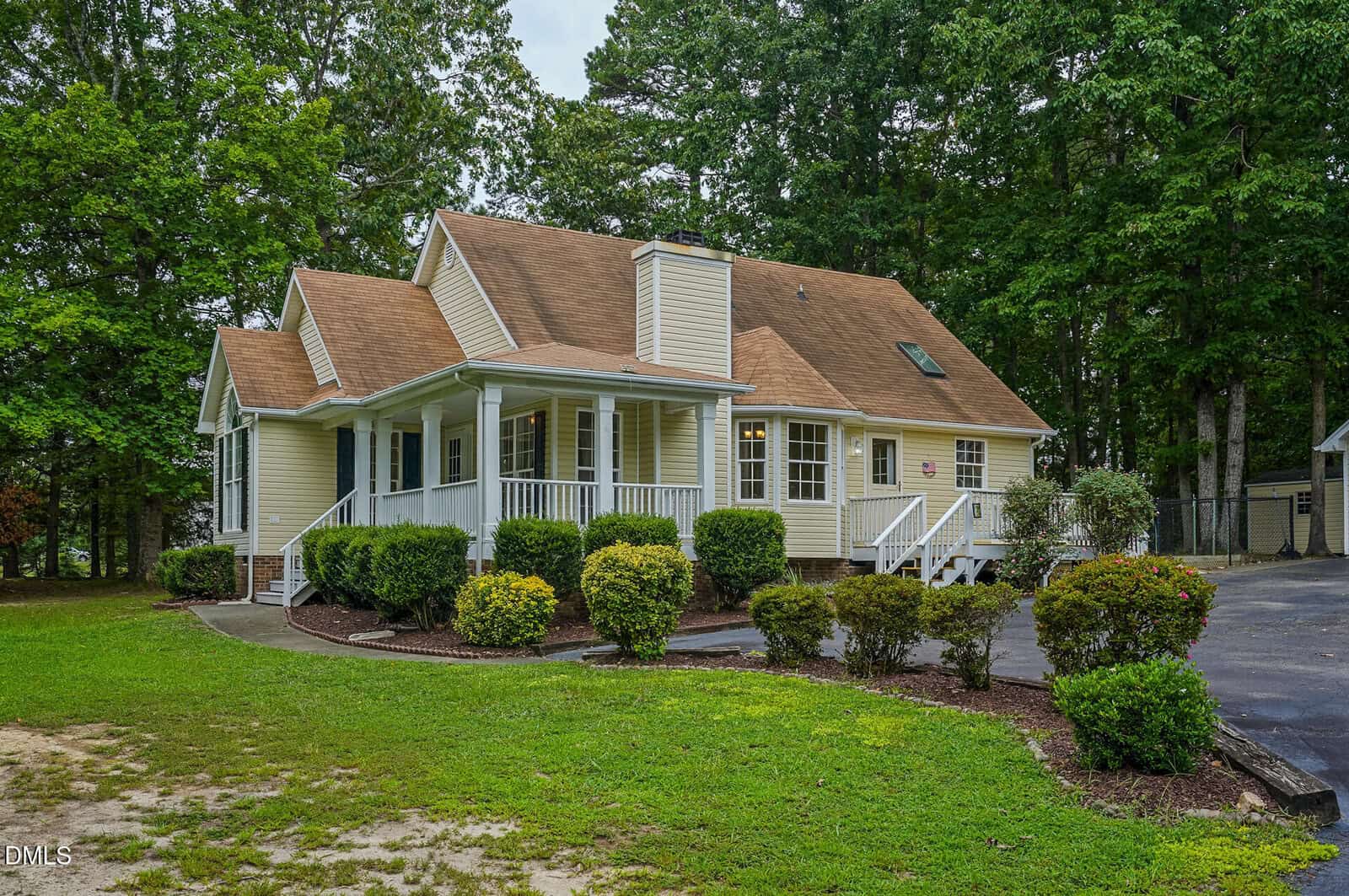 Yellow house with a brown roof, white trim, and a covered porch, surrounded by green shrubs and trees. A paved driveway curves in front of the home.