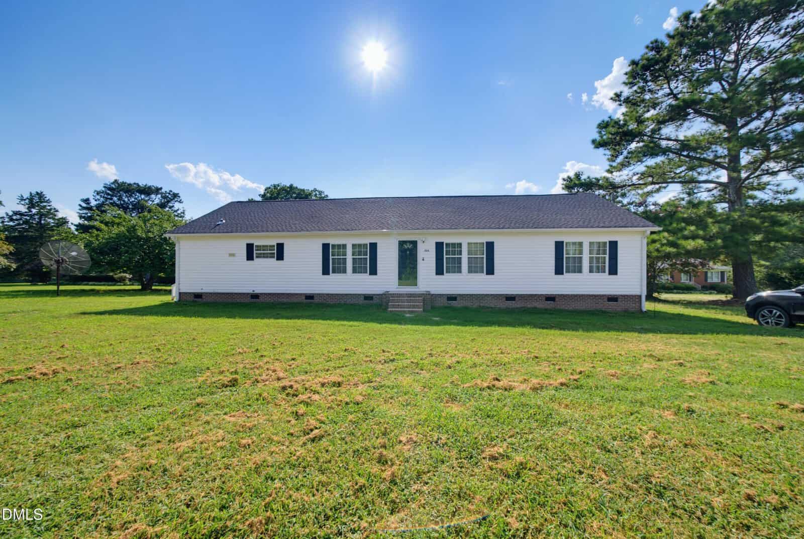A white single-story house with a dark roof sits on a large grassy yard under a bright sun and clear blue sky at 523 County Line Road in Manson, with a few trees nearby and a car partially visible on the right.