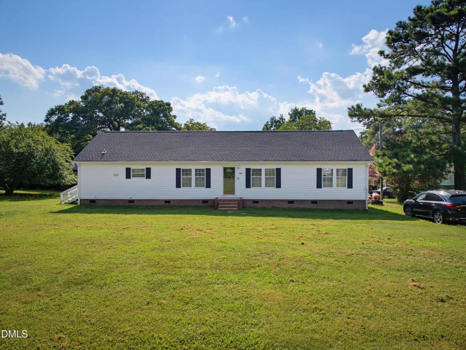 Single-story white house with black shutters and a dark roof, set on a large green lawn with a few trees and a black car parked to the right. The sky is bright and partly cloudy.