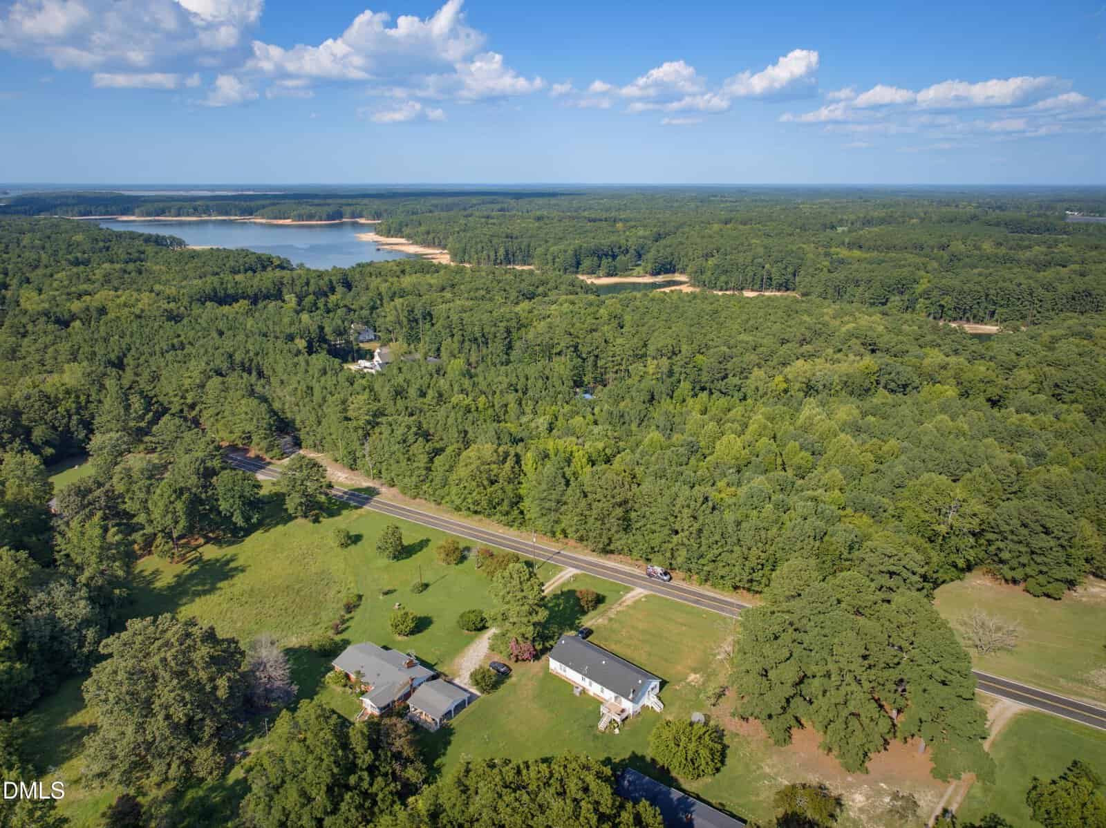 Aerial view of a rural area with houses, a road, dense green forest, and a lake in the background under a blue sky with scattered clouds.