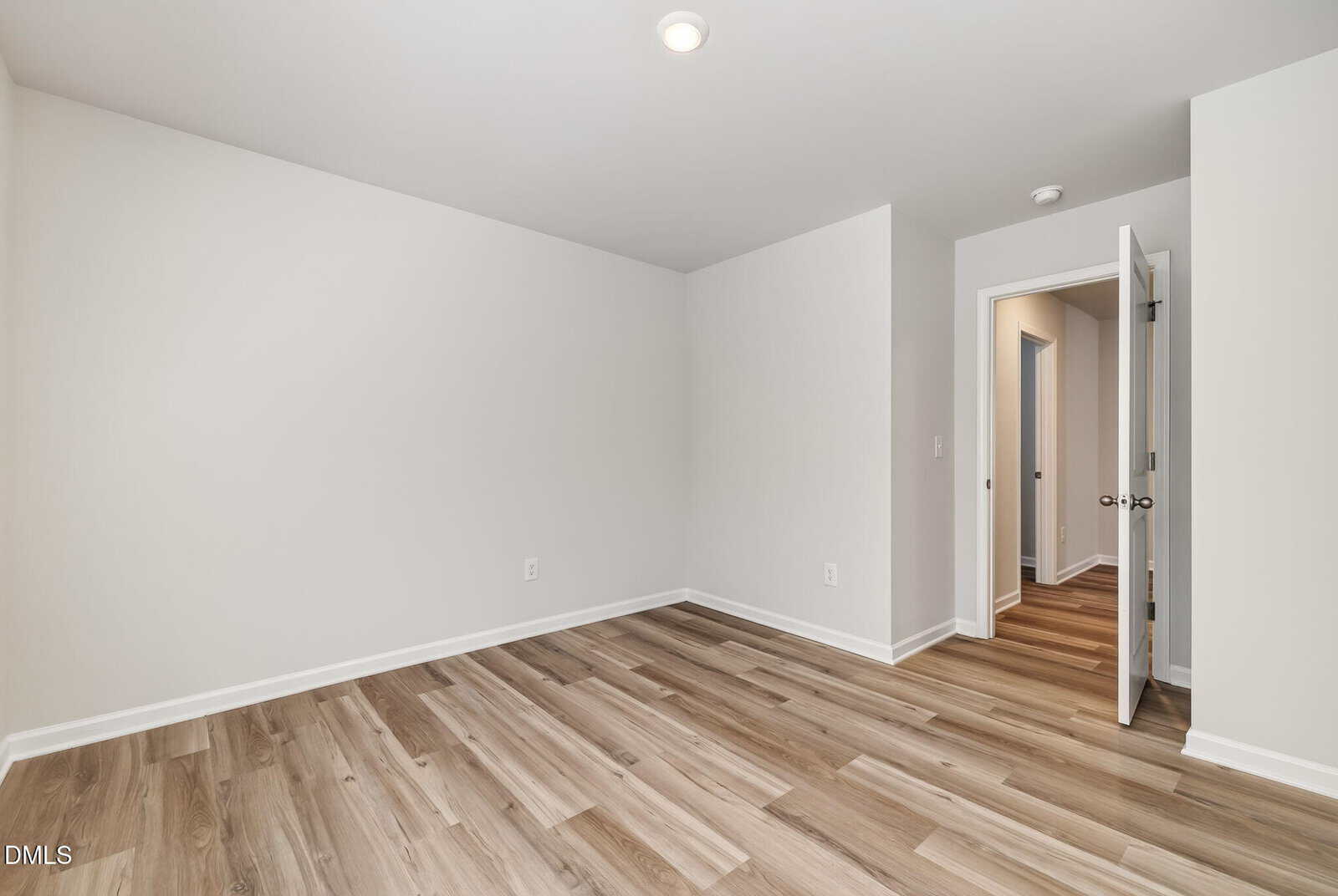 Empty room with light-colored walls, wood-look flooring, a ceiling light, and an open door leading to a hallway with similar flooring. The space is bright and modern with minimal features.