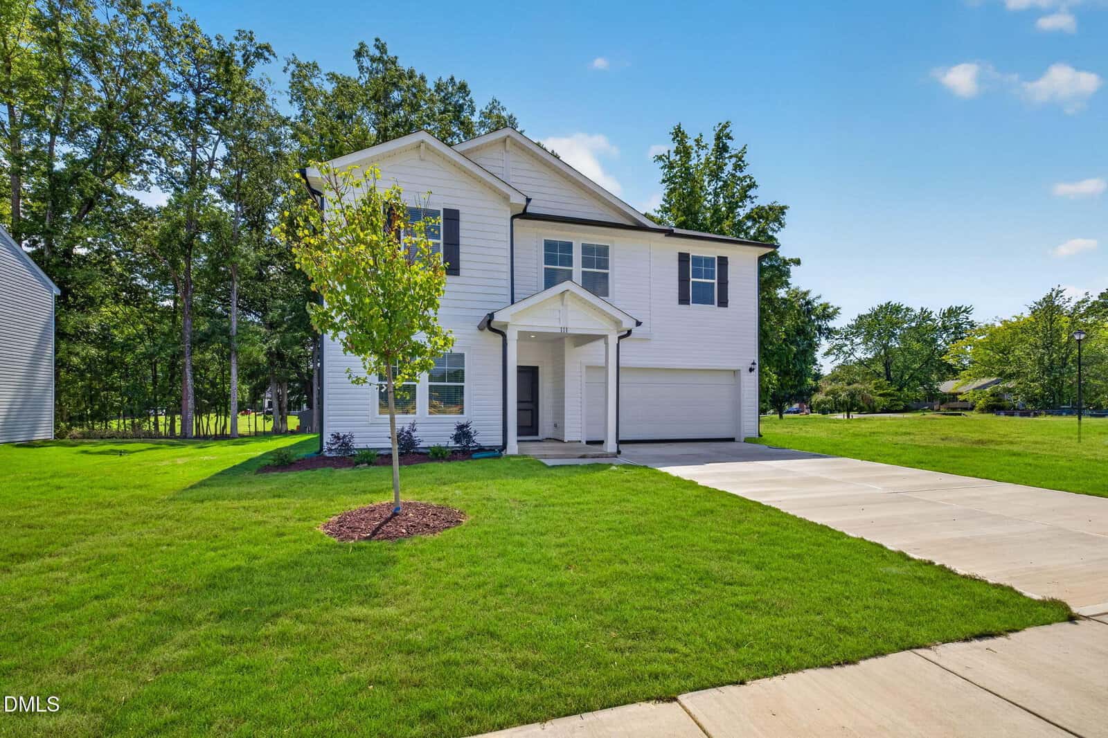 A modern two-story white house with black trim, a covered entryway, a double garage, and a concrete driveway. Green grass and a young tree are in the front yard, with trees and a blue sky in the background.