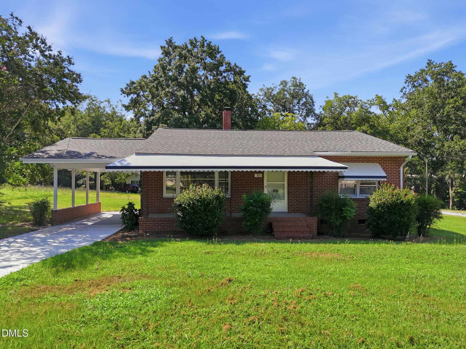 Charming single-story brick house at 311 W Mason Street, Franklinton, featuring an awning over the front windows, a covered carport on the left, a lush green lawn, and trees in the background under a blue sky.