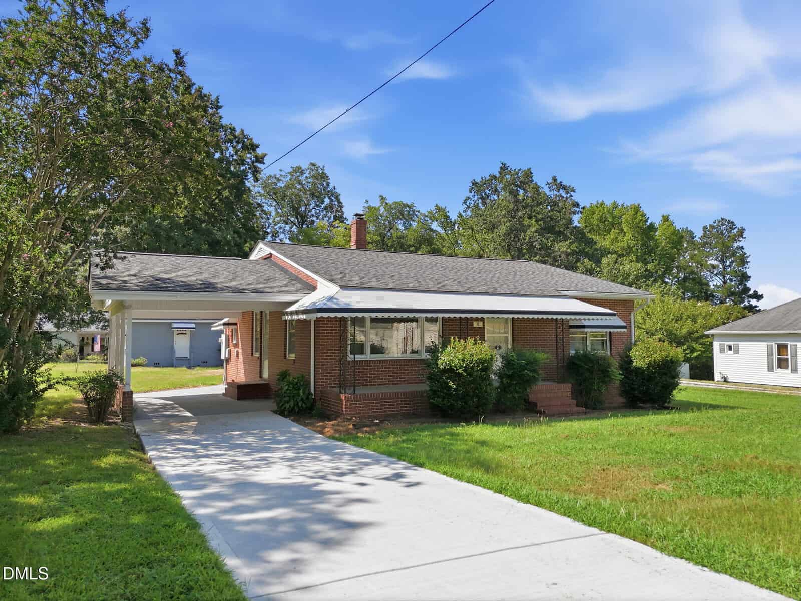 Brick single-story house with a gray roof, covered carport, front porch, bushes, and a concrete driveway, set amid a green lawn and trees under a blue sky.