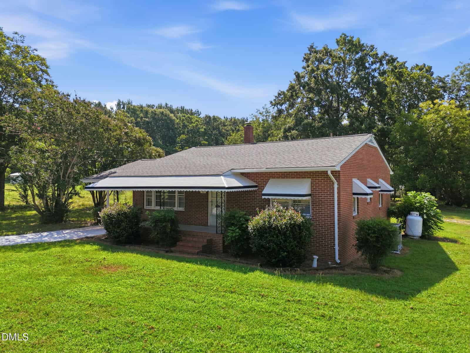 A single-story red brick house with a gray roof, white awnings, and a covered front porch sits on a grassy lawn with shrubs and trees nearby under a blue sky.