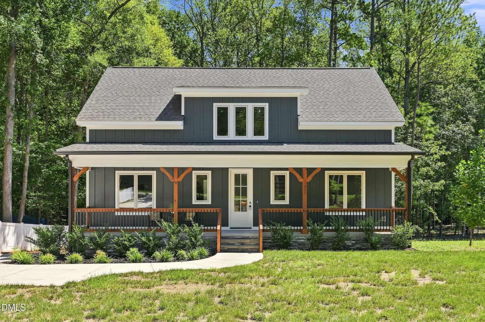 A modern gray house at 117 Pool Rock Shores Lane, Henderson, featuring a covered front porch with wooden railings, white trim, and a charming dormer window, is surrounded by trees and a lush green lawn on a sunny day.