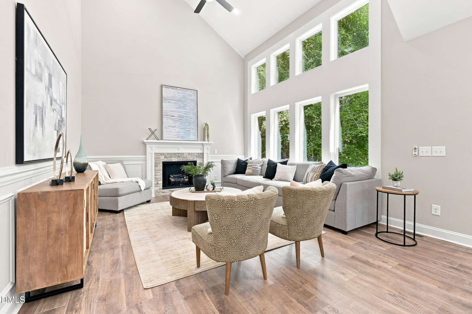 Modern living room with light gray walls, large windows, a sectional sofa, wooden coffee table, two patterned chairs, fireplace, artwork, plants, and wood flooring. Bright natural light fills the space.