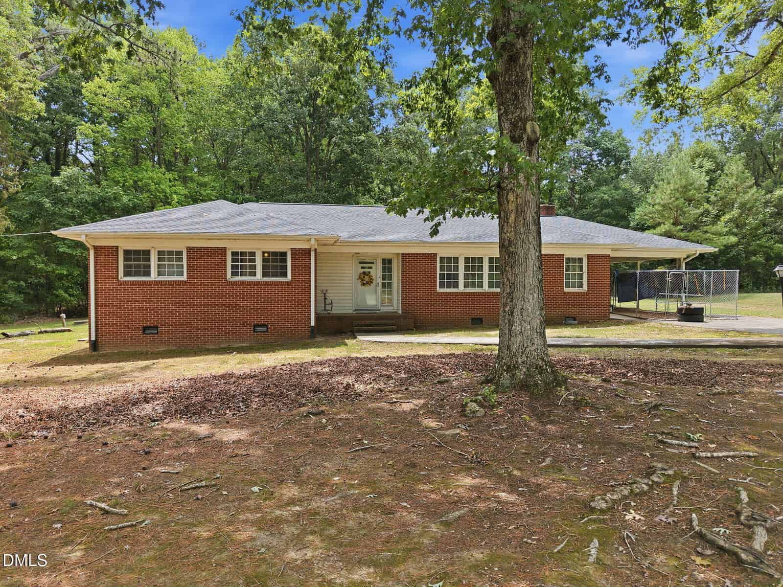 Charming single-story red brick house with white trim, a gray shingle roof, and a welcoming front porch located at 1136 Mountain Creek Road, Oxford. Surrounded by trees and a large yard with scattered leaves and branches, this property also features a chain-link fence visible on the right.
